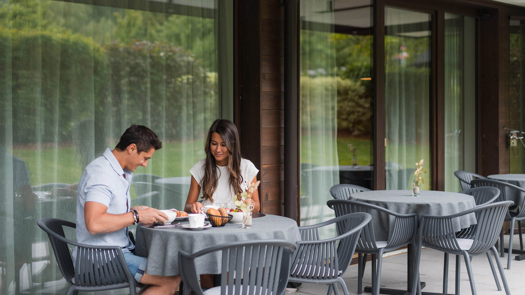 A man and woman sit at an outdoor café table, smiling, with breakfast and drinks in front of them. - Hotel Oberwiesen