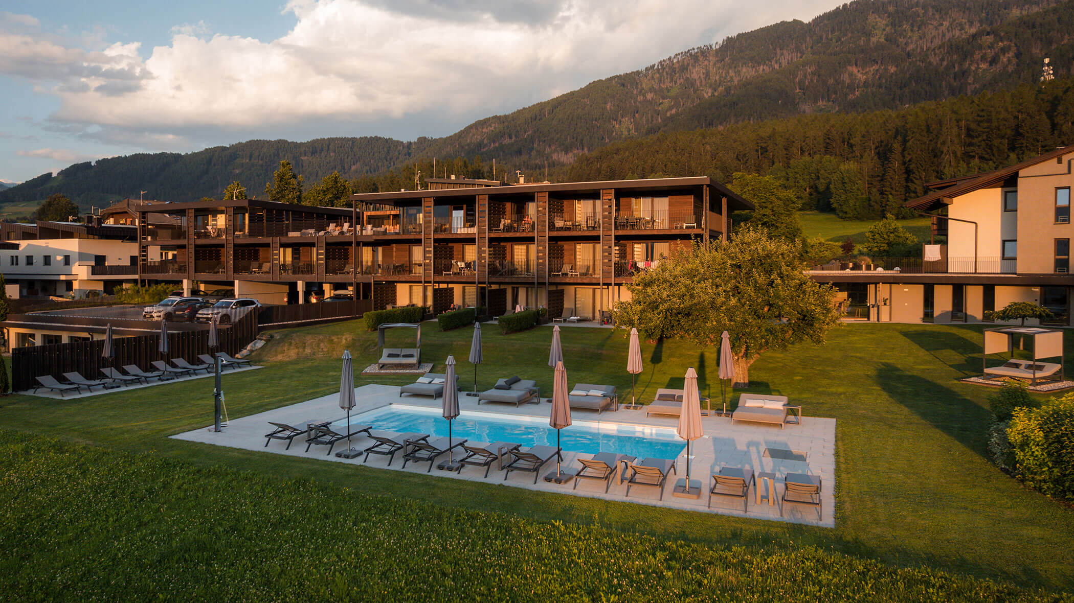 Modern hotel with wooden balconies, outdoor pool, lounge chairs, and umbrellas, set against forested hills. - Hotel Oberwiesen