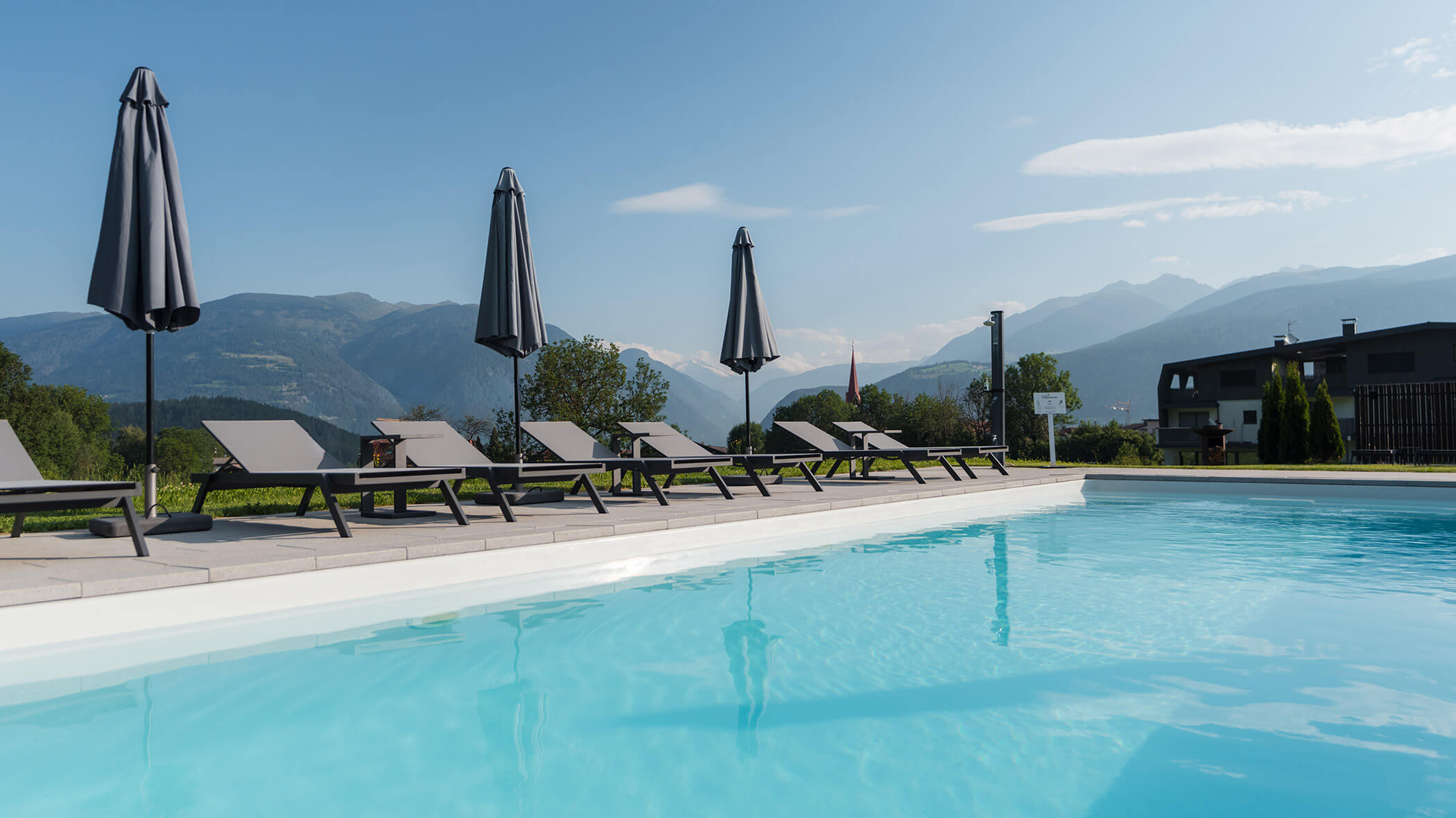 Empty lounge chairs with closed umbrellas line a pool, mountains visible in the background under blue sky. - Hotel Oberwiesen
