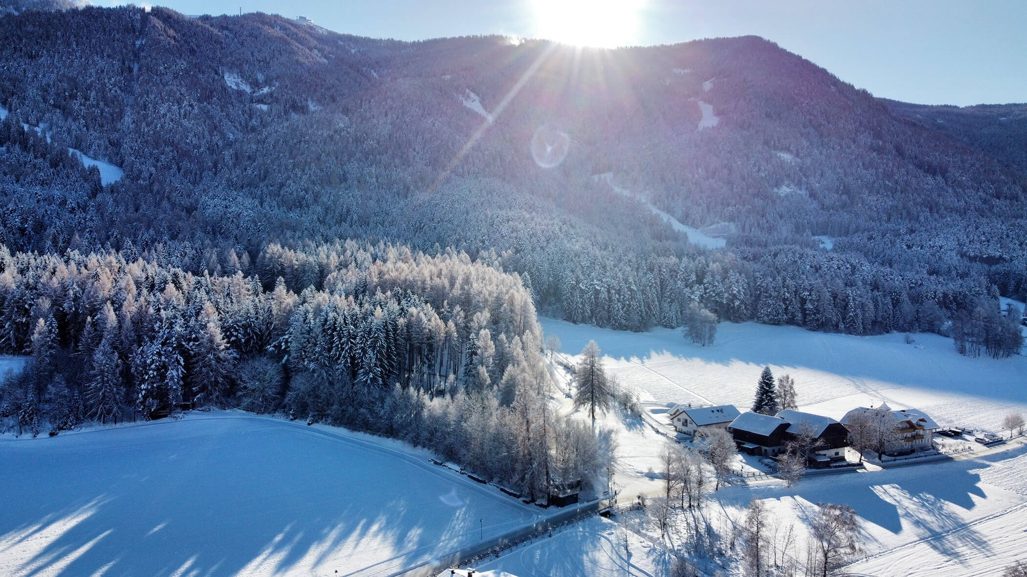 A snowy landscape with trees, houses, and mountains under a bright sun in a clear blue sky. - Hotel Oberwiesen