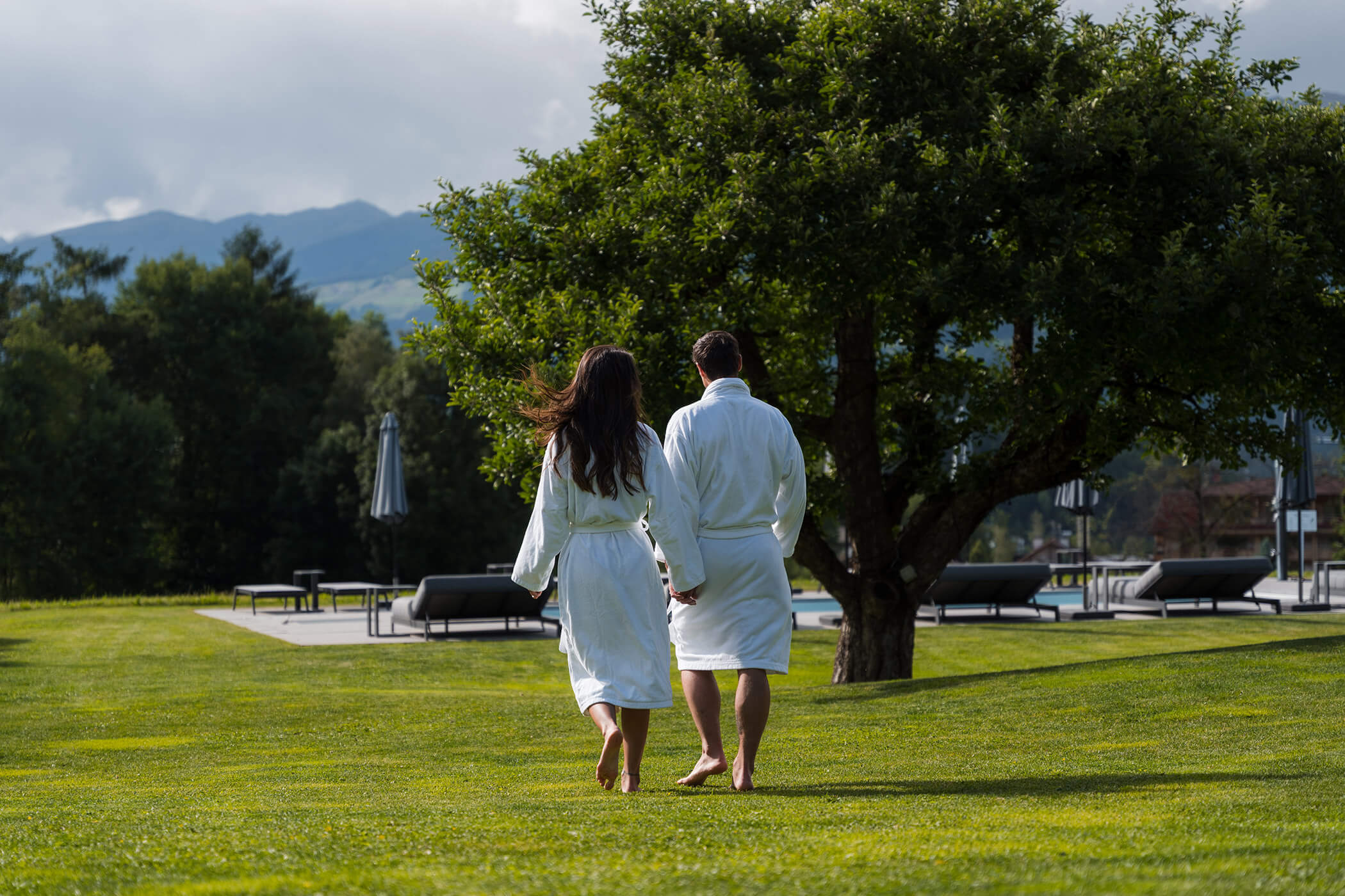 A couple in white robes walks hand in hand on grass toward a tree, with lounge chairs and mountains in the background. - Hotel Oberwiesen