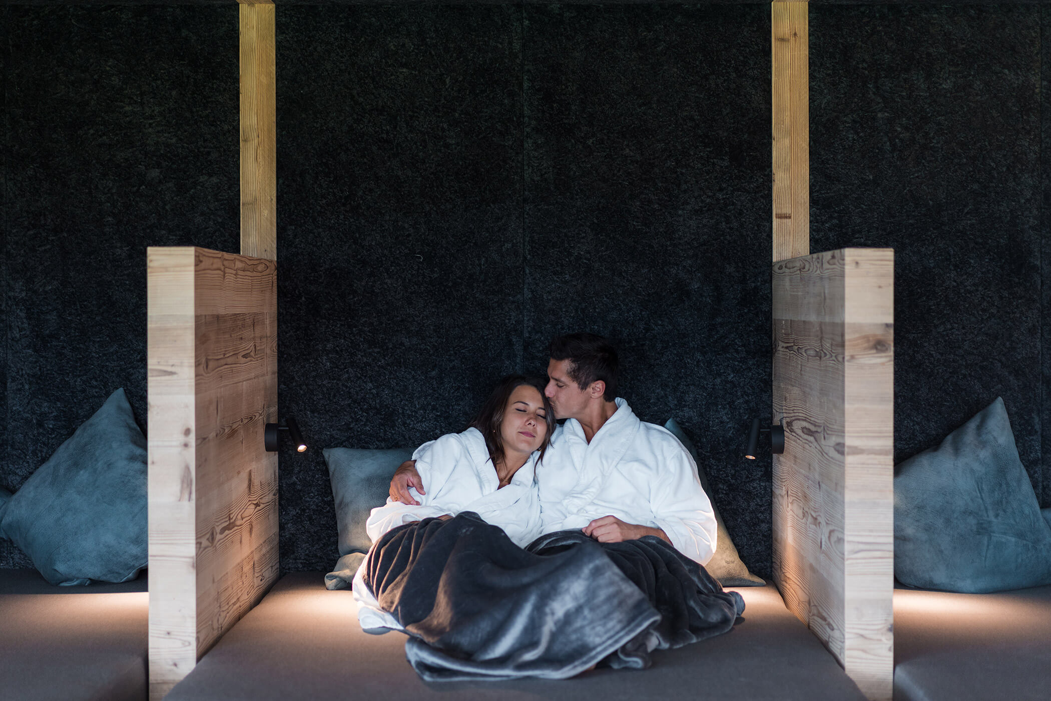 A couple in white robes cuddles under a gray blanket, relaxing on a cozy bed with wooden dividers. - Hotel Oberwiesen