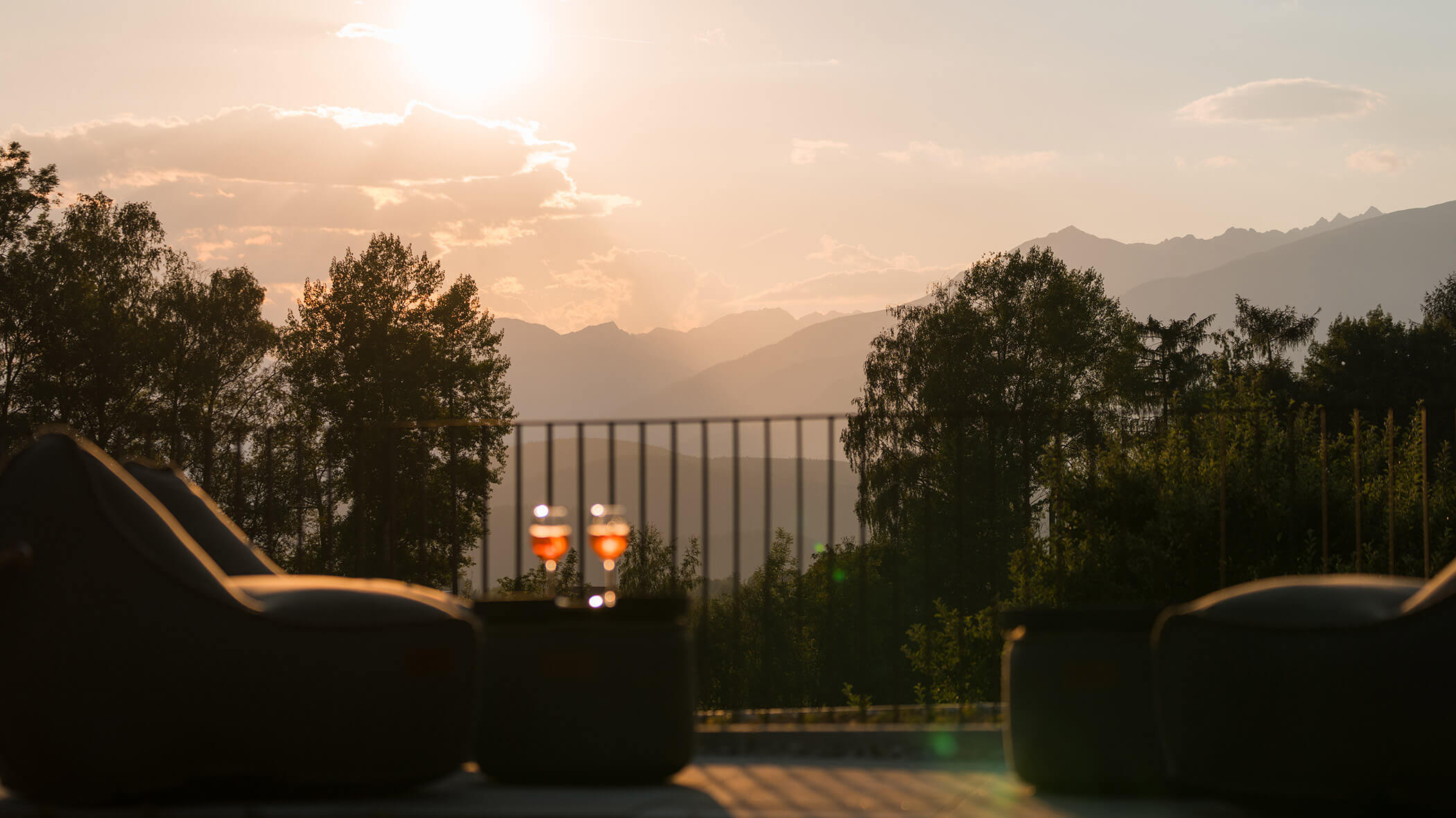 Two glasses of rosé on a table, with trees and mountains at sunset in the background. - Hotel Oberwiesen