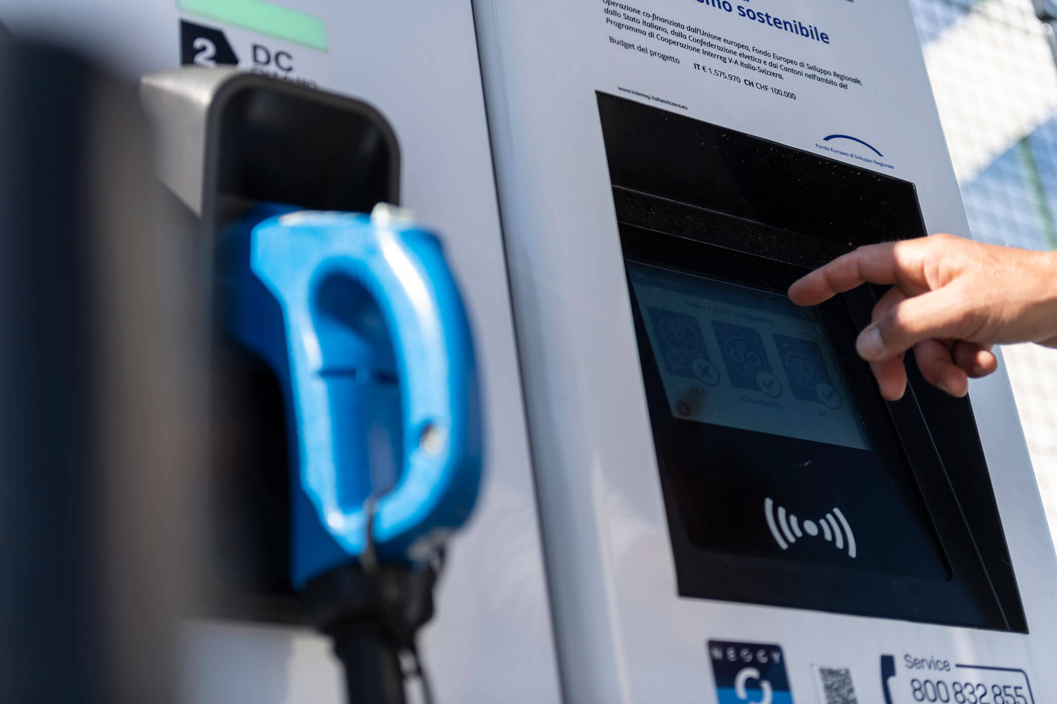 Eine Hand berührt den Bildschirm einer Ladestation für Elektrofahrzeuge, neben der sich ein blauer Stecker befindet. - Hotel Oberwiesen