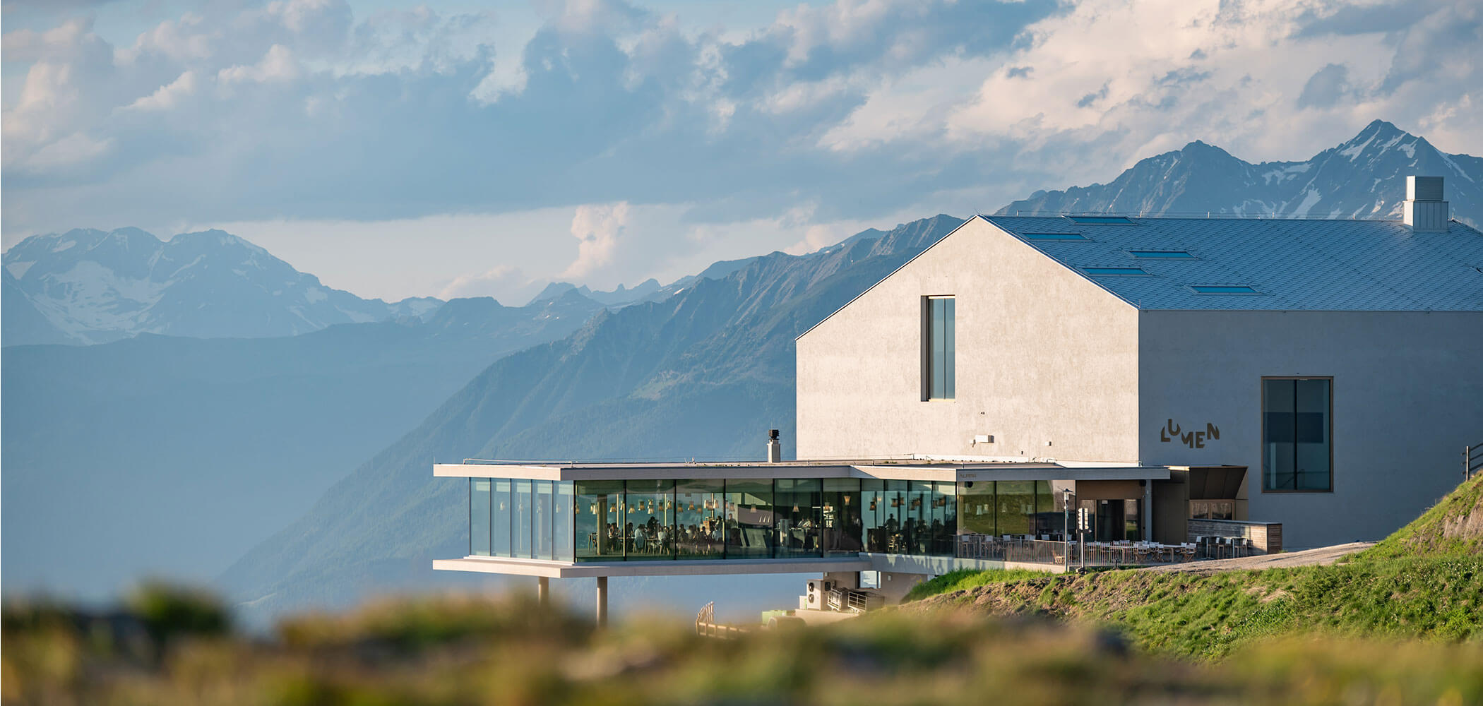 Un edificio moderno con pareti in vetro si affaccia sulle montagne sotto un cielo nuvoloso, con tavoli disposti all’aperto in un patio. - Hotel Oberwiesen