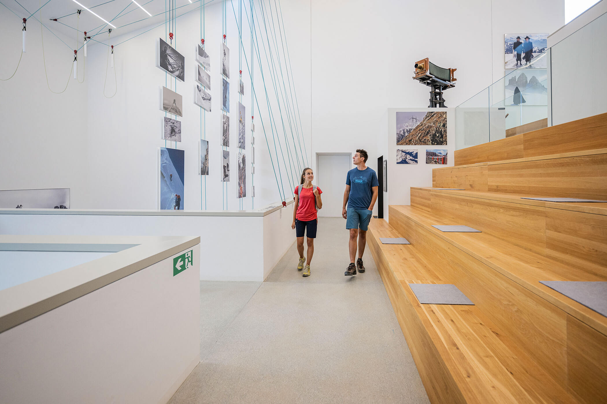 Due persone camminano accanto a tribune di legno in una galleria moderna dove sono appese, appese a dei fili, delle foto di montagne. - Hotel Oberwiesen