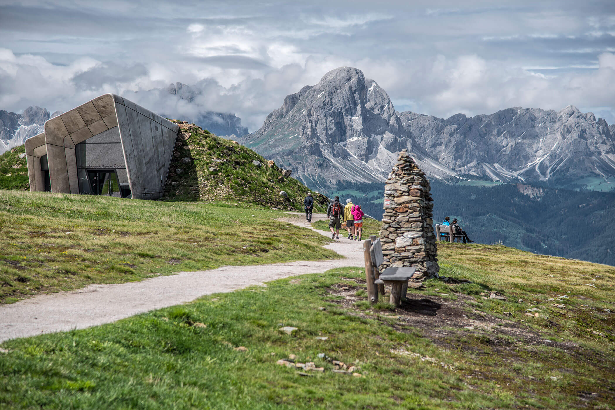 Gli escursionisti percorrono un sentiero di montagna vicino a un moderno edificio in pietra e a un cumulo di pietre, con le cime delle montagne sullo sfondo. - Hotel Oberwiesen