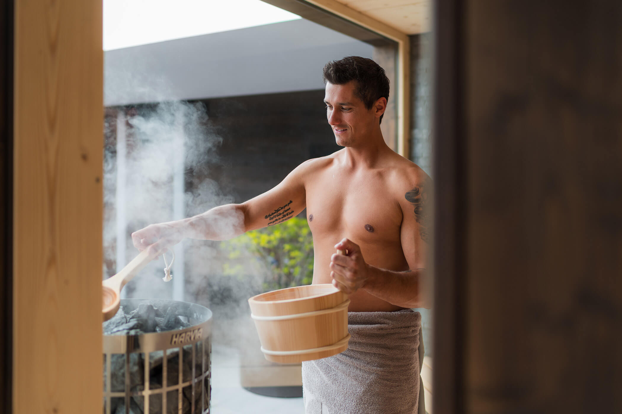 Ein Mann ohne Hemd, nur mit einem Handtuch bekleidet, gießt Wasser auf die Saunasteine und erzeugt so Dampf in einer Holzsauna. - Hotel Oberwiesen