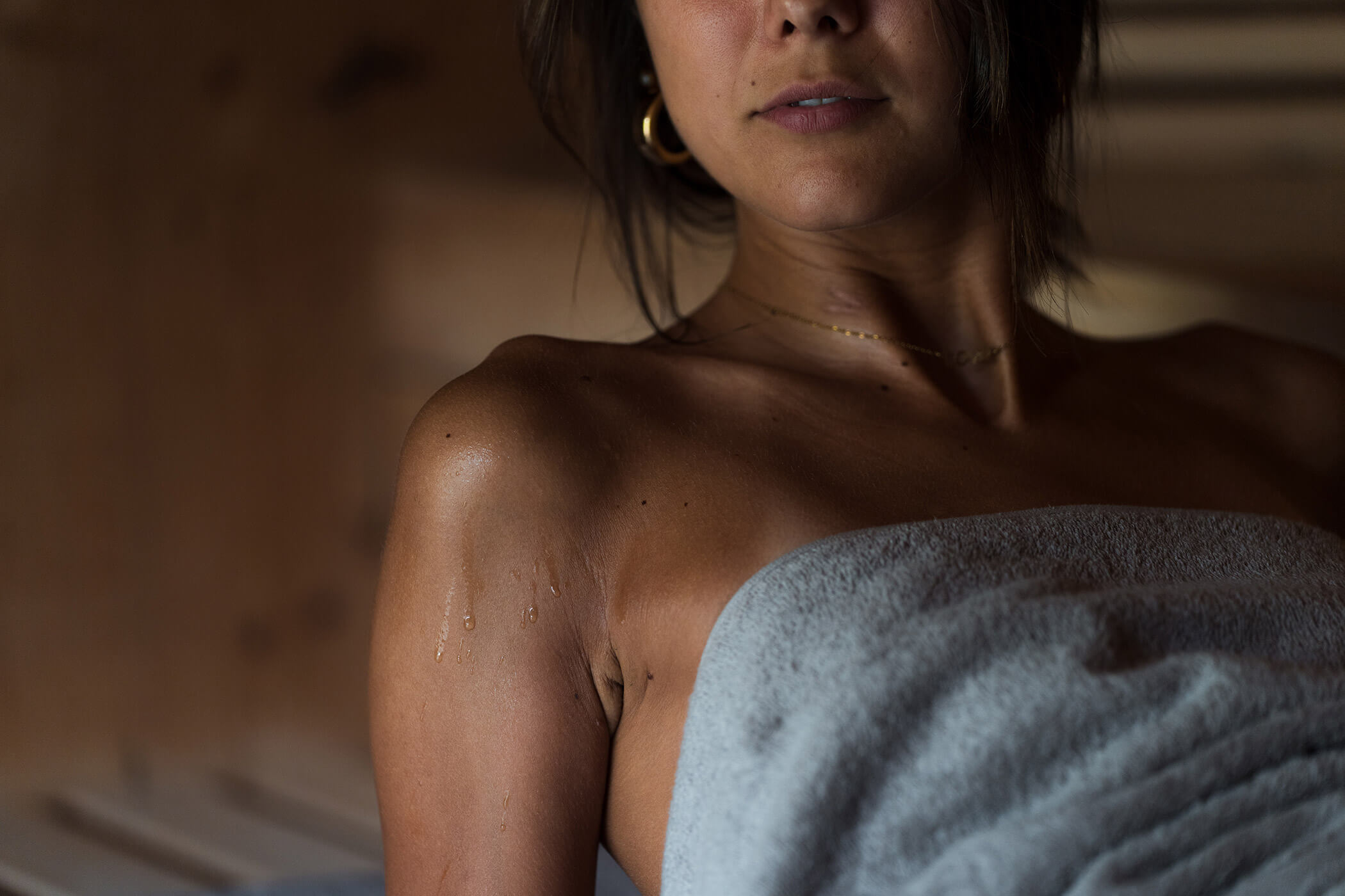 Eine Frau mit feuchter Haut sitzt in einer Sauna, in ein Handtuch gehüllt, mit Wassertropfen auf der Schulter. - Hotel Oberwiesen