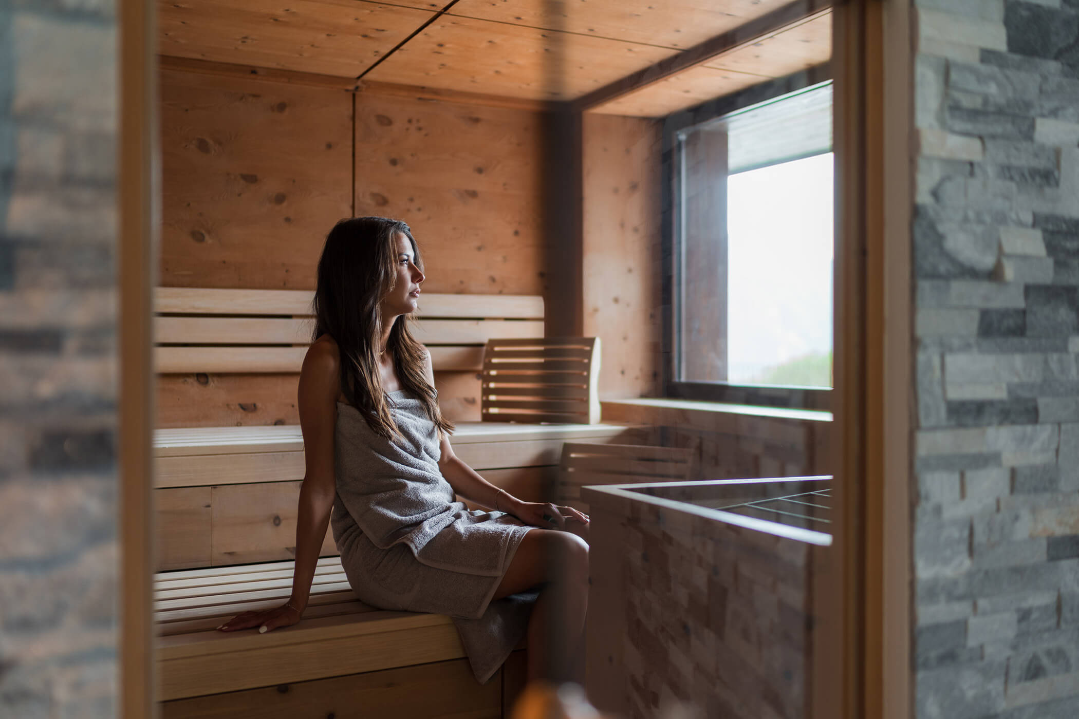 Eine Frau in einem Handtuch sitzt auf einer Holzbank in einer Sauna und schaut aus dem Fenster. - Hotel Oberwiesen