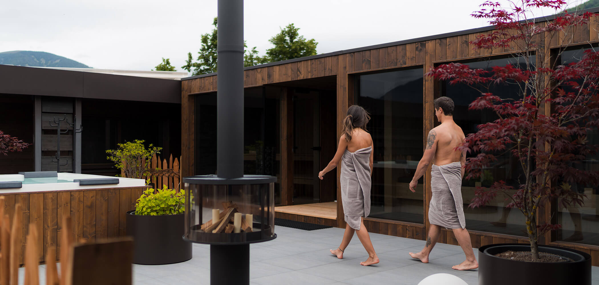 Zwei Menschen in Handtüchern spazieren auf einer modernen Außenterrasse in der Nähe eines Holzgebäudes mit Pflanzen und einem Kamin. - Hotel Oberwiesen