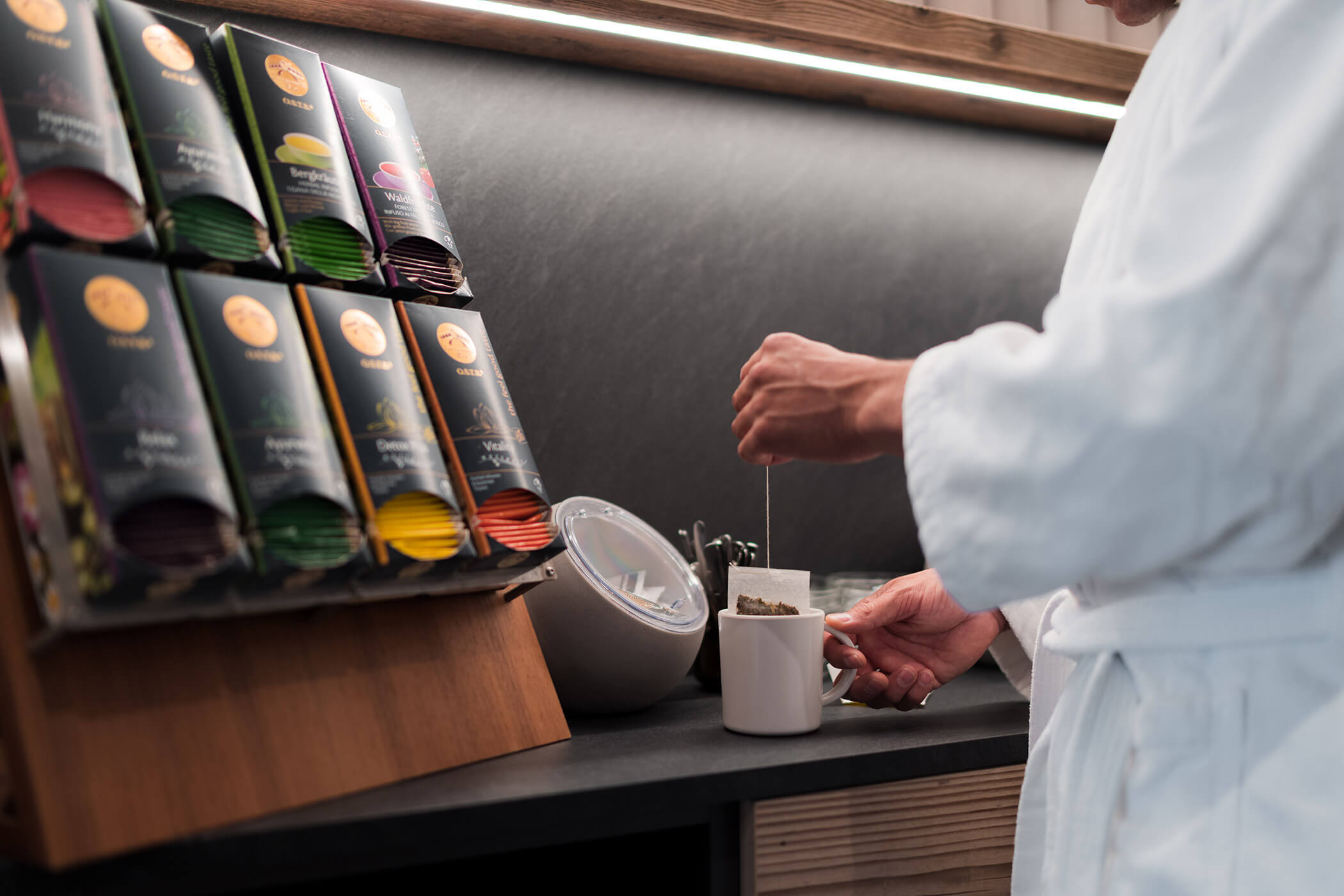 Eine Person in einem weißen Gewand, die an einer Theke mit einer Teeauswahl und Zuckerpäckchen Tee zubereitet. - Hotel Oberwiesen
