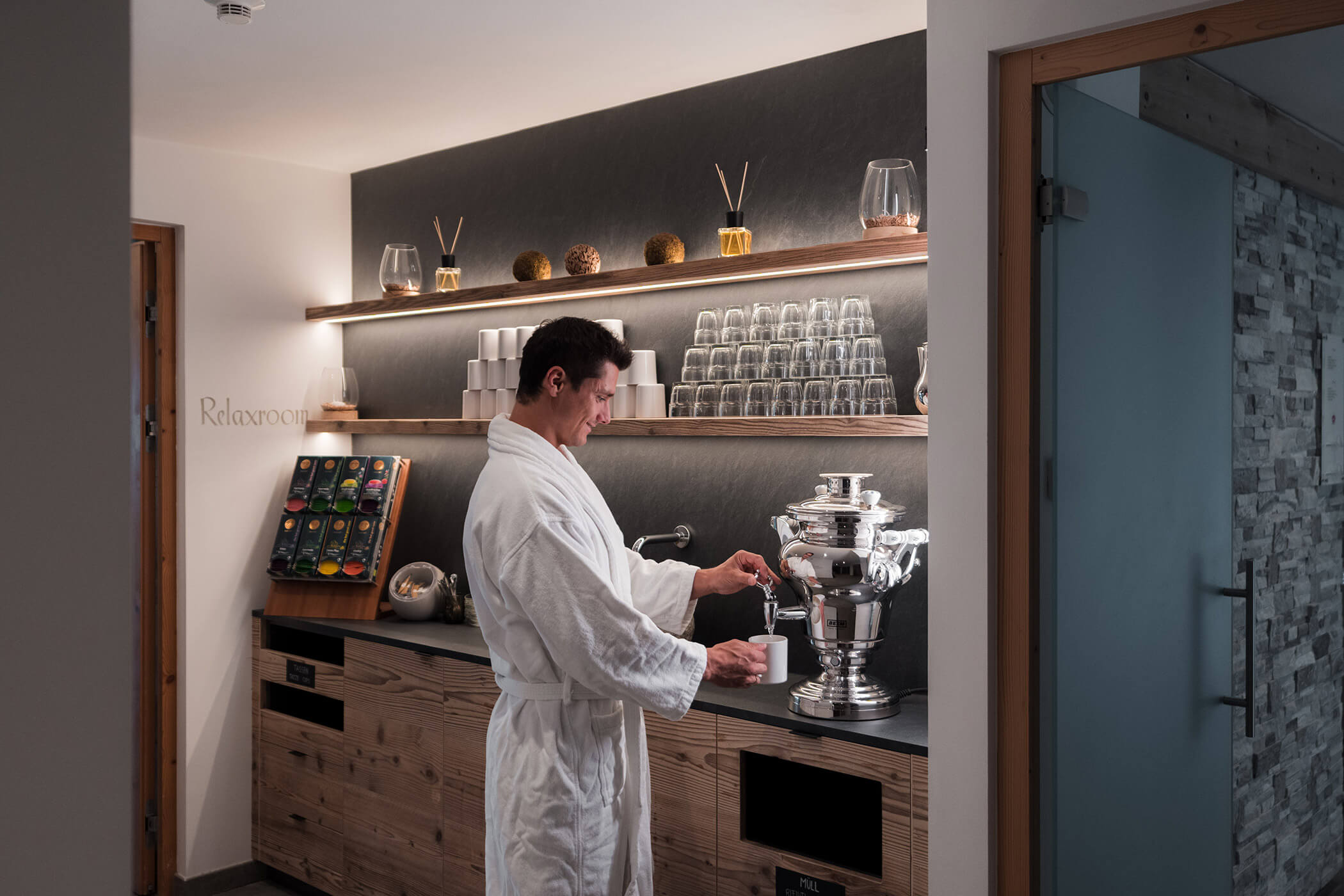 Ein Mann in einem weißen Gewand schenkt Tee aus einem Samowar in einem modernen, gemütlichen Ruheraum mit Regalen und Glasgeschirr ein. - Hotel Oberwiesen