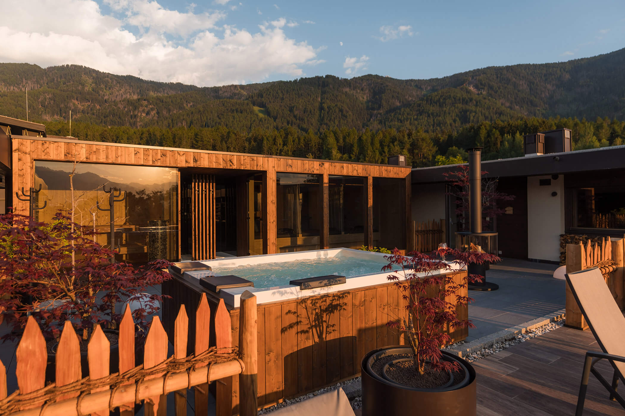 Whirlpool im Freien auf einer Holzterrasse mit Blick auf die Berge, umgeben von einem Zaun und Topfpflanzen. - Hotel Oberwiesen
