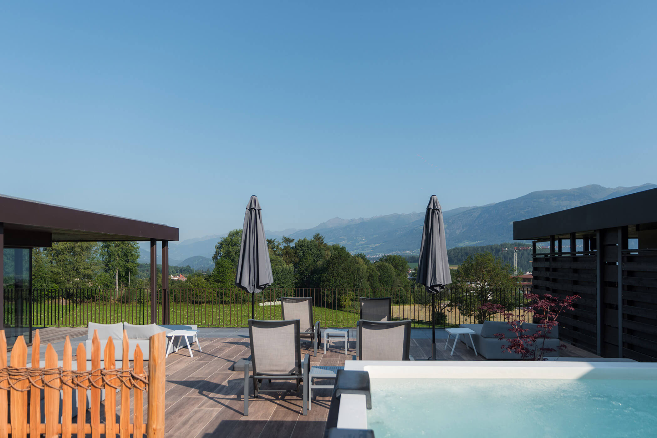 Die Terrasse mit Liegestühlen, Sonnenschirmen und Whirlpool bietet einen Blick auf die Rasenfläche und die fernen Berge unter blauem Himmel. - Hotel Oberwiesen