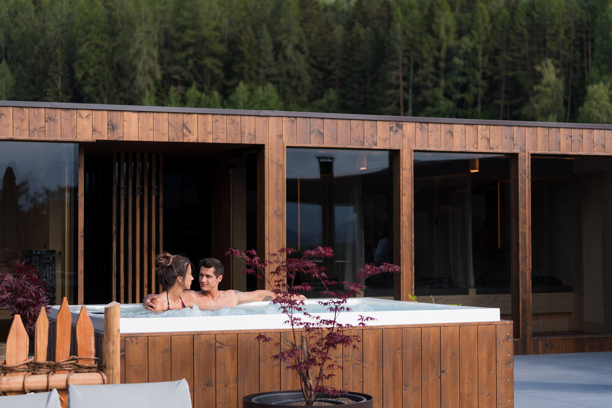 Ein Paar entspannt sich in einem Whirlpool im Freien neben einem Holzgebäude, im Hintergrund ist ein Wald zu sehen. - Hotel Oberwiesen