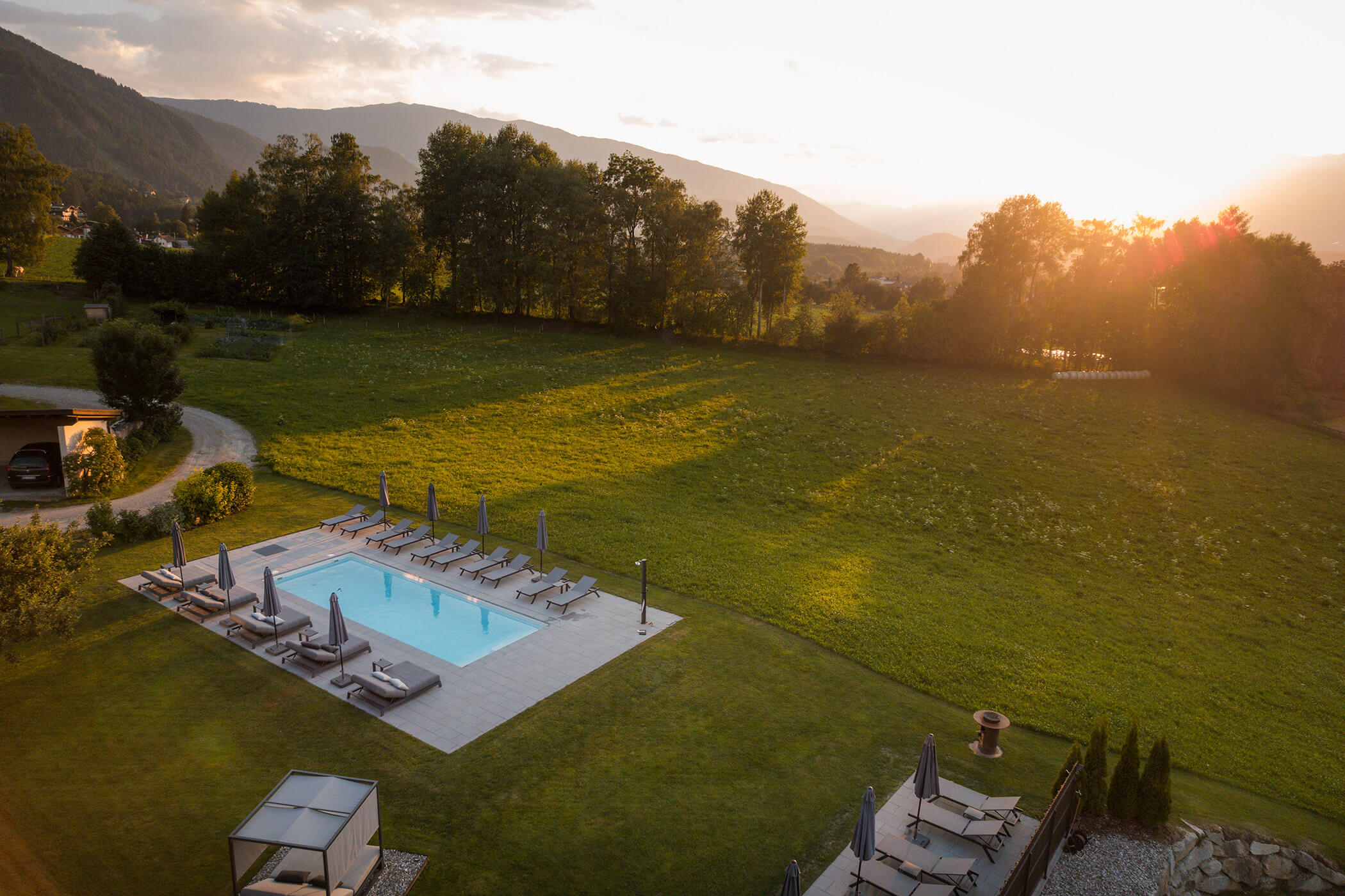 Ein rechteckiges Freibad mit Liegestühlen liegt bei Sonnenuntergang neben einer offenen, grünen Wiese. - Hotel Oberwiesen