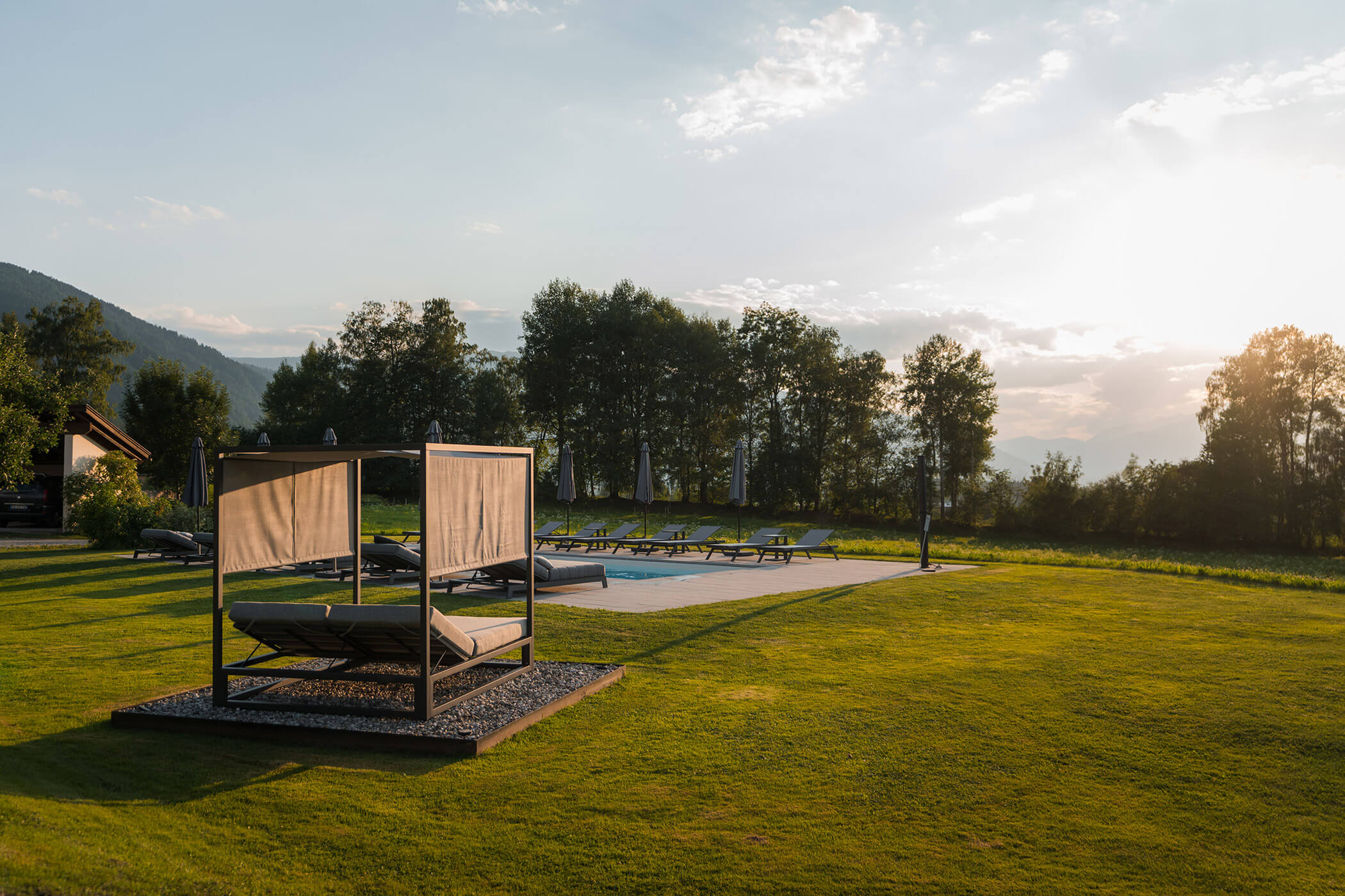 Sonnenuntergang über einem Rasen mit einer Gartenhütte, einem Pool, Liegestühlen und Bäumen im Hintergrund. - Hotel Oberwiesen