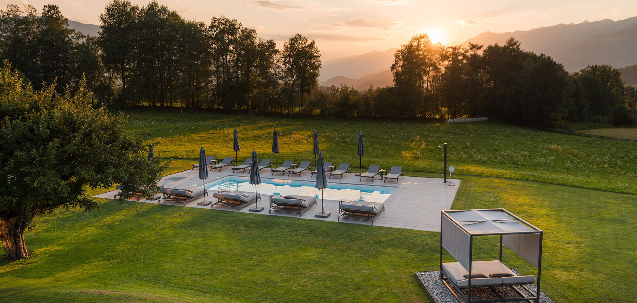 Außenpool mit Liegestühlen und Sonnenschirmen, umgeben von grüner Rasenfläche und Bäumen bei Sonnenuntergang. - Hotel Oberwiesen
