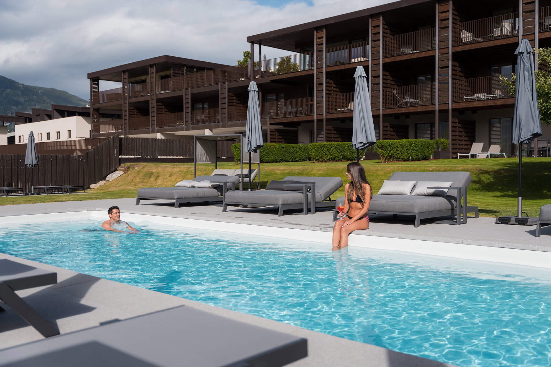 Eine Frau sitzt am Pool, während ein Mann schwimmt; im Hintergrund sind Liegestühle und Wohnungen zu sehen. - Hotel Oberwiesen
