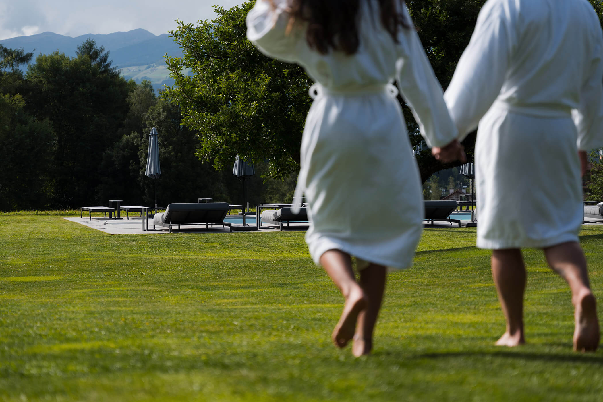 Zwei Menschen in weißen Gewändern gehen barfuß über das Gras auf Liegestühle und Sonnenschirme in einem sonnigen Außenbereich zu. - Hotel Oberwiesen