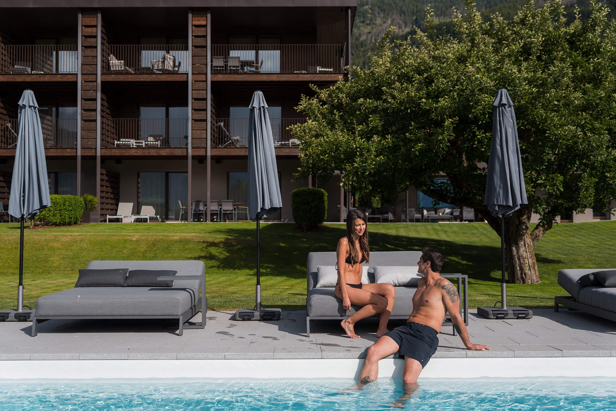 Ein Mann und eine Frau in Badekleidung entspannen sich am Pool, umgeben von Liegestühlen und Sonnenschirmen, im Hintergrund ist ein modernes Gebäude zu sehen. - Hotel Oberwiesen