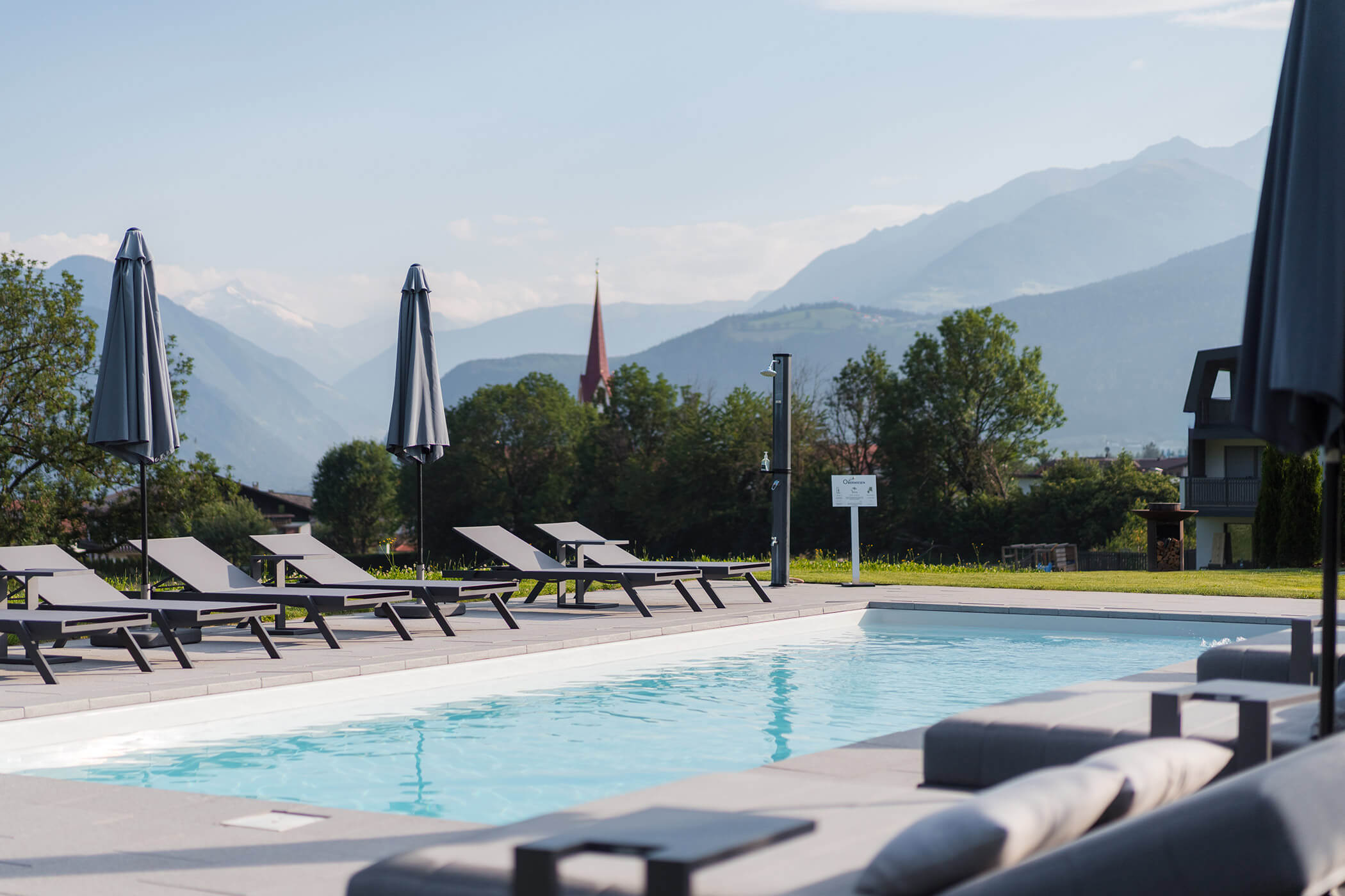 Moderner Pool mit Liegestühlen und Sonnenschirmen, umgeben von Bergen und Grünflächen unter strahlend blauem Himmel. - Hotel Oberwiesen