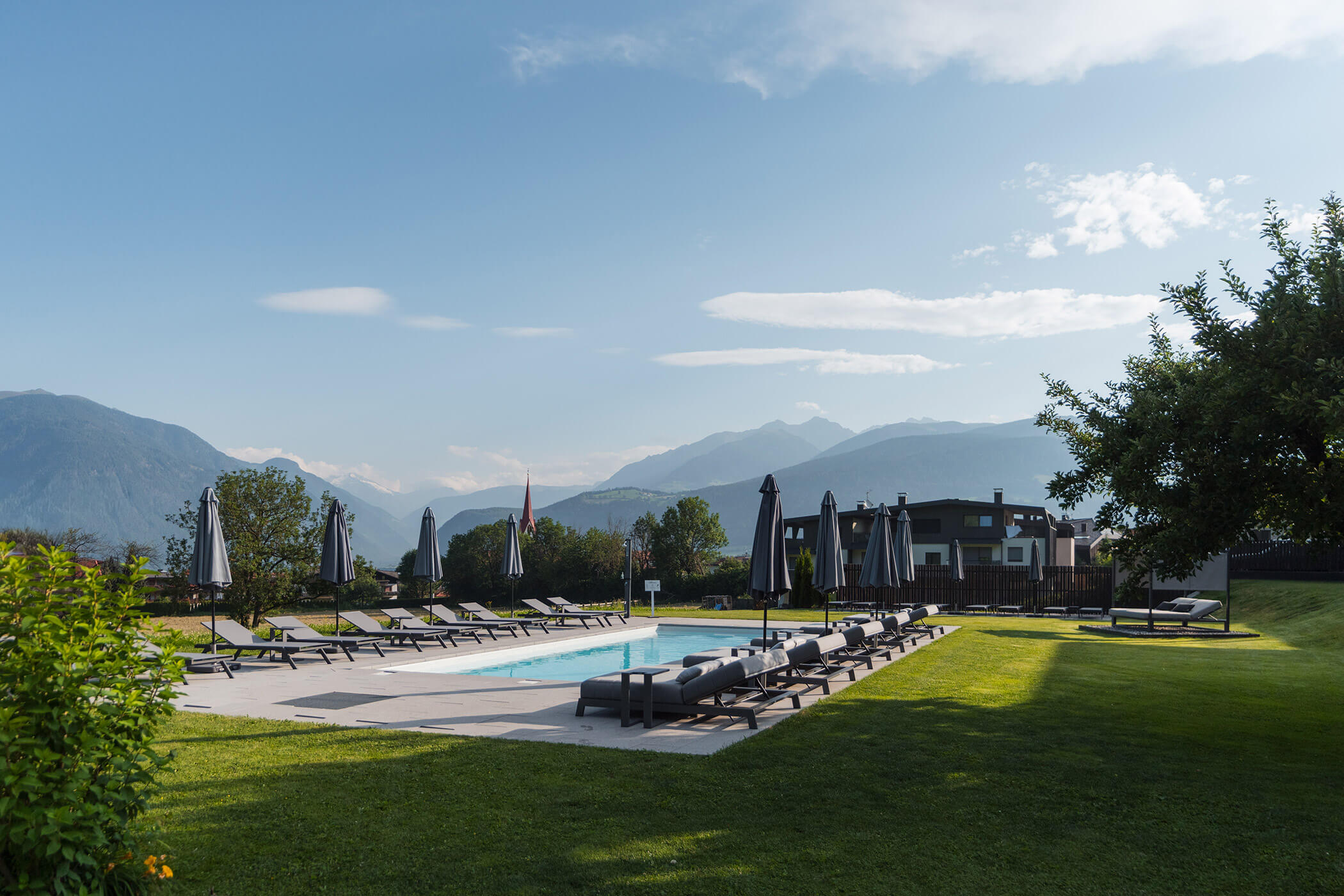Außenpool mit Liegestühlen und Sonnenschirmen, vor der Kulisse der Berge und einer Rasenfläche unter blauem Himmel. - Hotel Oberwiesen