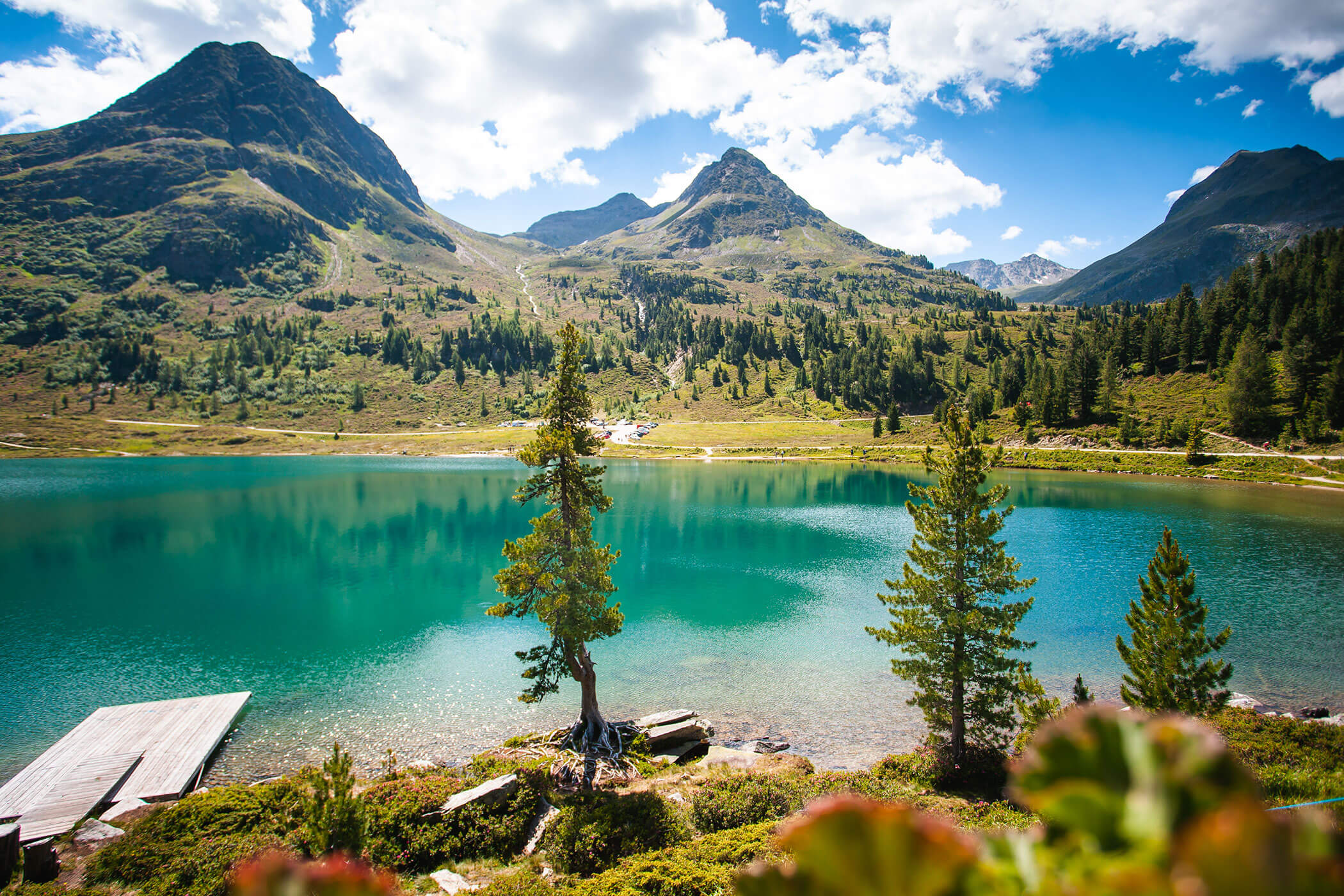 Ein klarer, türkisfarbener See mit einem Steg, Kiefern und Bergen unter einem teilweise bewölkten Himmel. - Hotel Oberwiesen