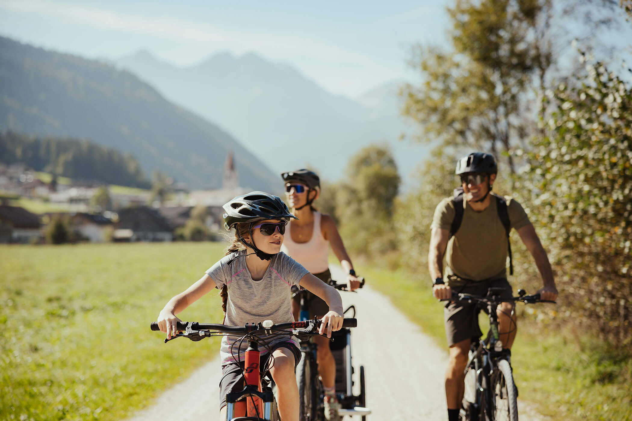 Drei Menschen fahren mit dem Fahrrad auf einem Feldweg durch eine sonnige, malerische Landschaft, im Hintergrund sind Berge zu sehen. - Hotel Oberwiesen