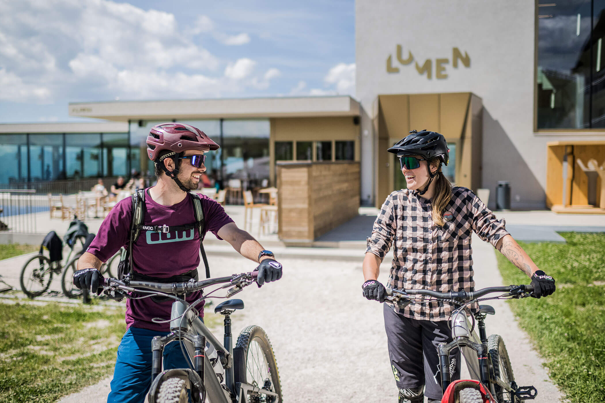 An einem sonnigen Tag lächeln sich zwei Menschen mit Fahrrädern vor einem modernen Gebäude mit der Aufschrift „LUMEN“ an. - Hotel Oberwiesen