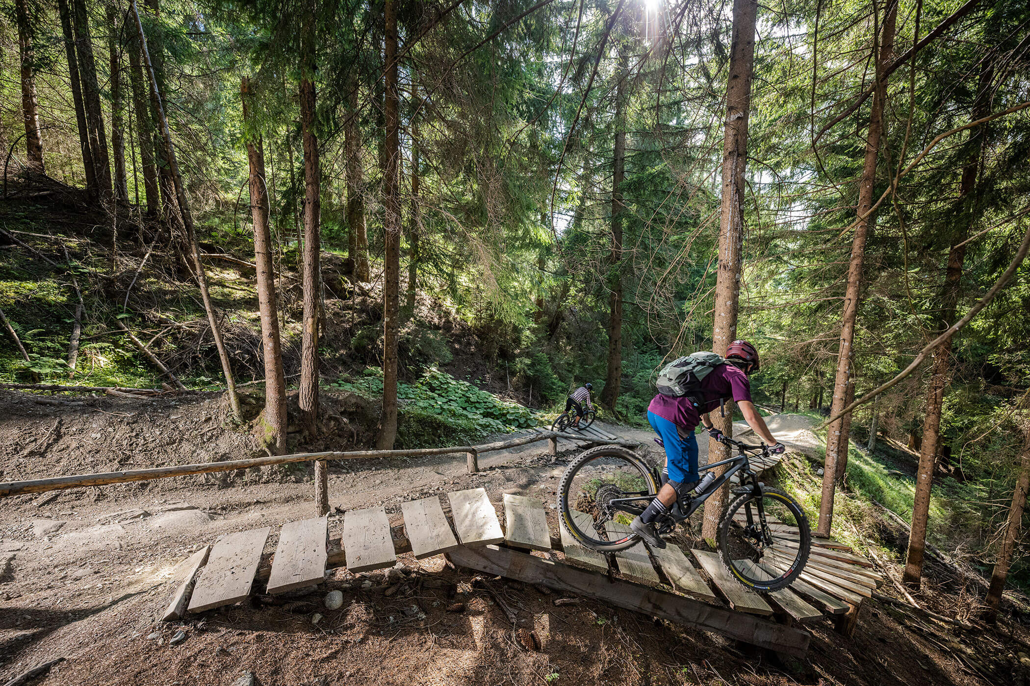 Eine Person fährt mit dem Mountainbike auf einem Holzpfad bergauf durch einen dichten, sonnendurchfluteten Wald. - Hotel Oberwiesen