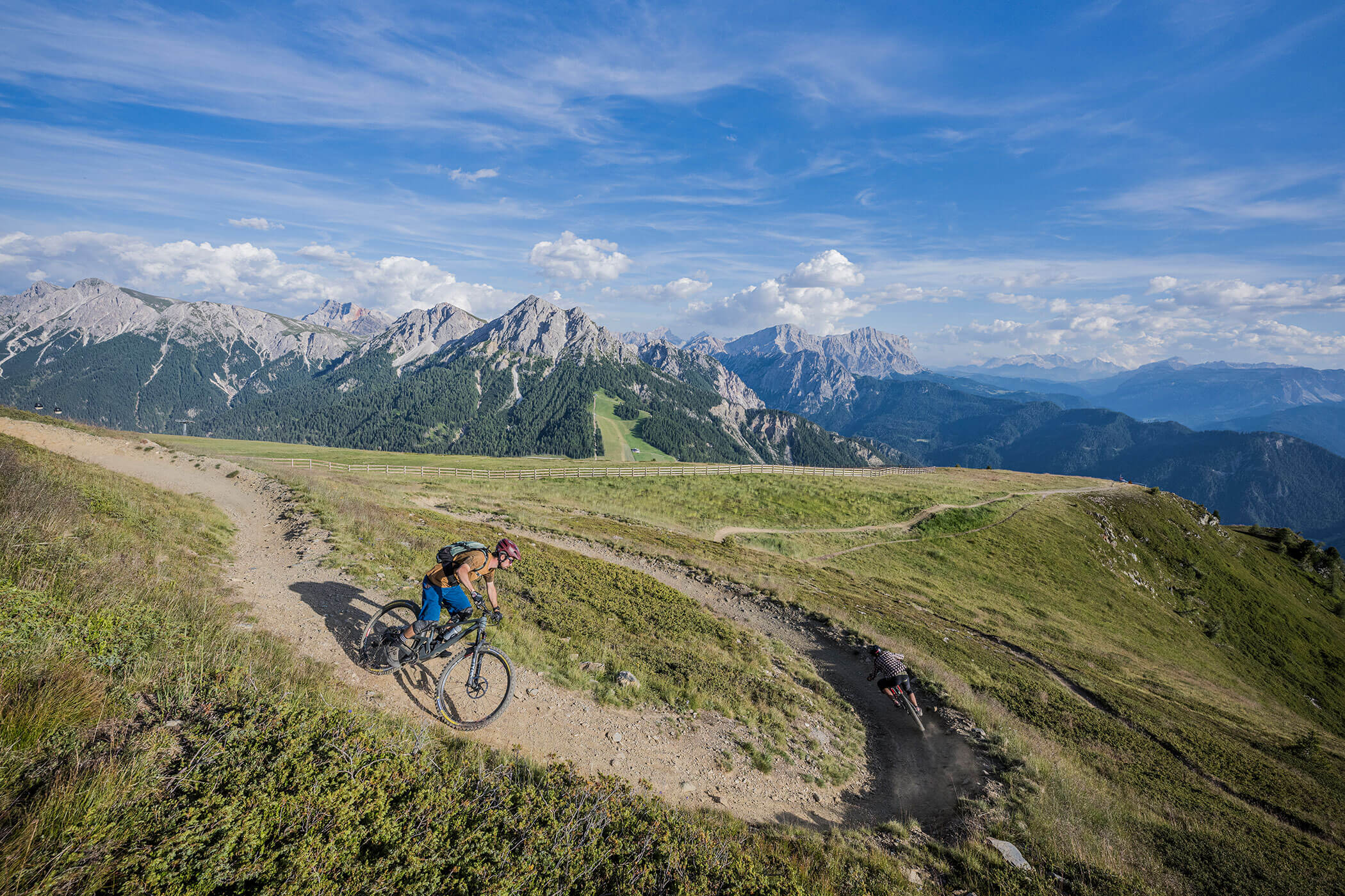 Mountainbiker fahren auf kurvenreichen Feldwegen an einem grasbewachsenen Alphang entlang, im Hintergrund ragen Berge empor. - Hotel Oberwiesen