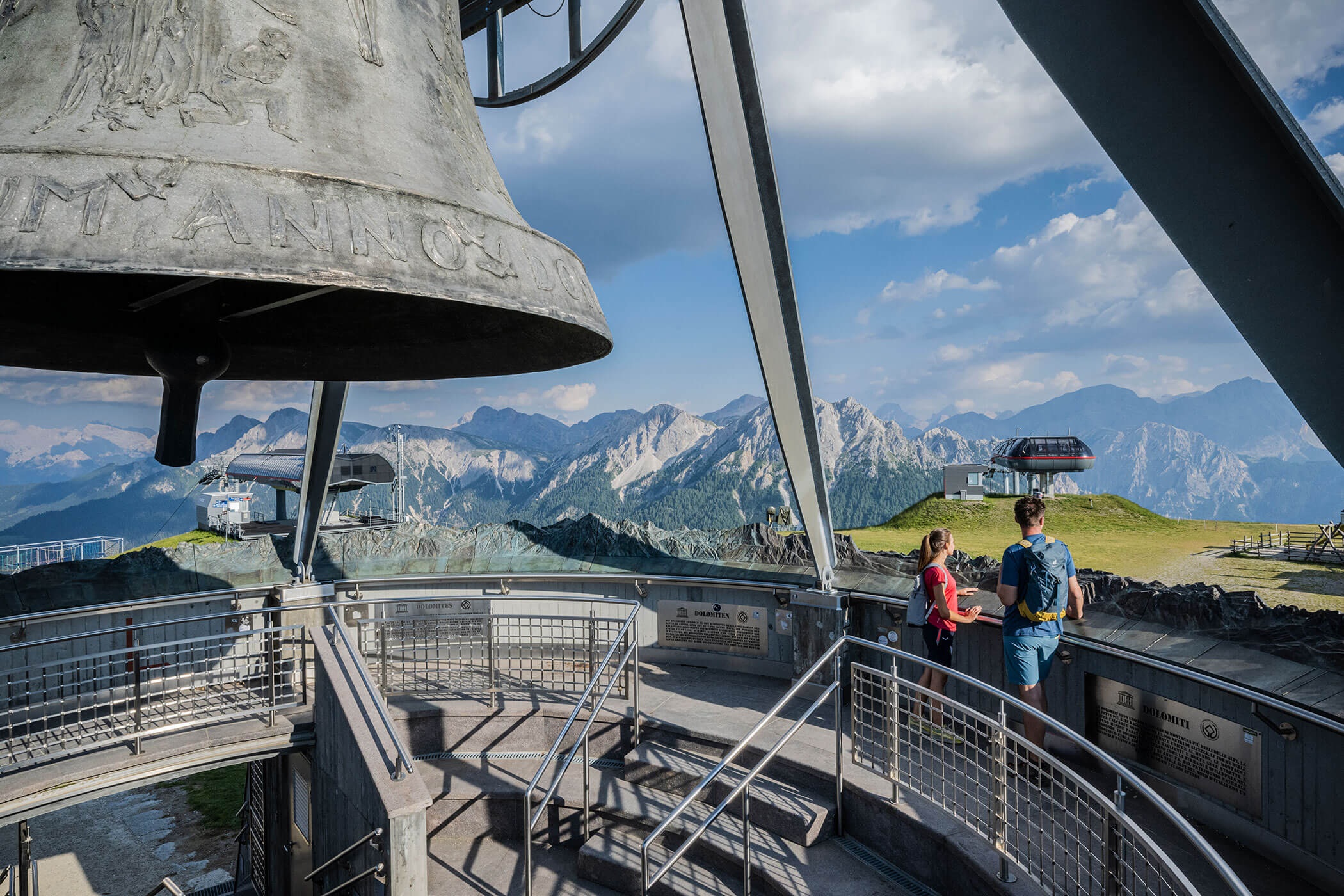 Zwei Menschen stehen auf einer Aussichtsplattform auf einem Berggipfel, im Hintergrund sind eine große Glocke und malerische Berge zu sehen. - Hotel Oberwiesen
