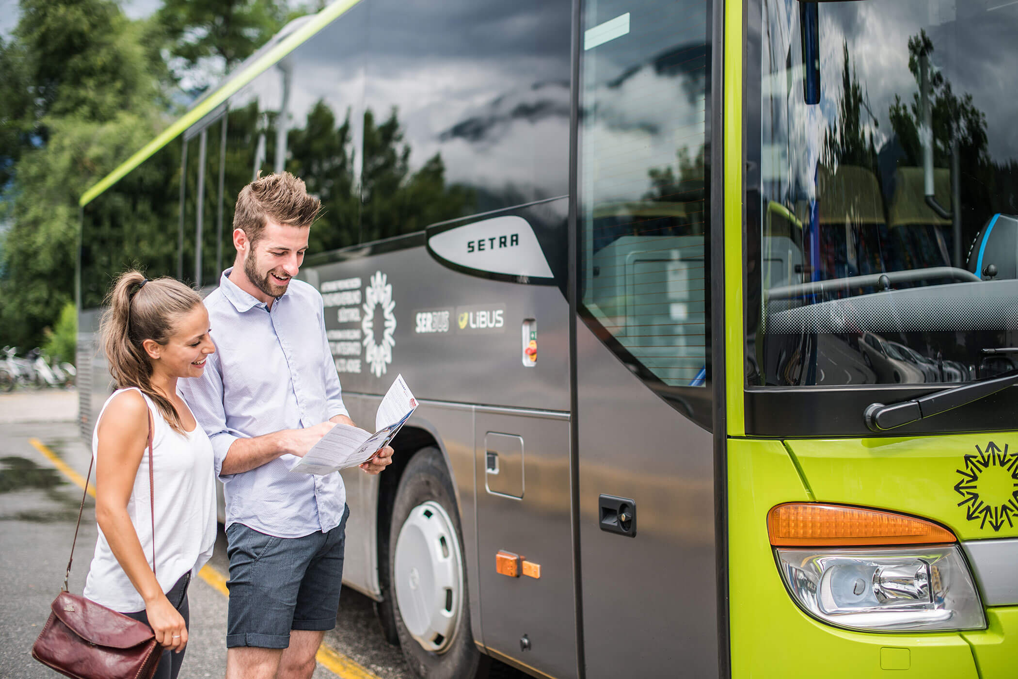 Ein Mann und eine Frau stehen neben einem grünen Bus, lächeln und schauen sich eine Karte oder eine Reisebroschüre an. - Hotel Oberwiesen