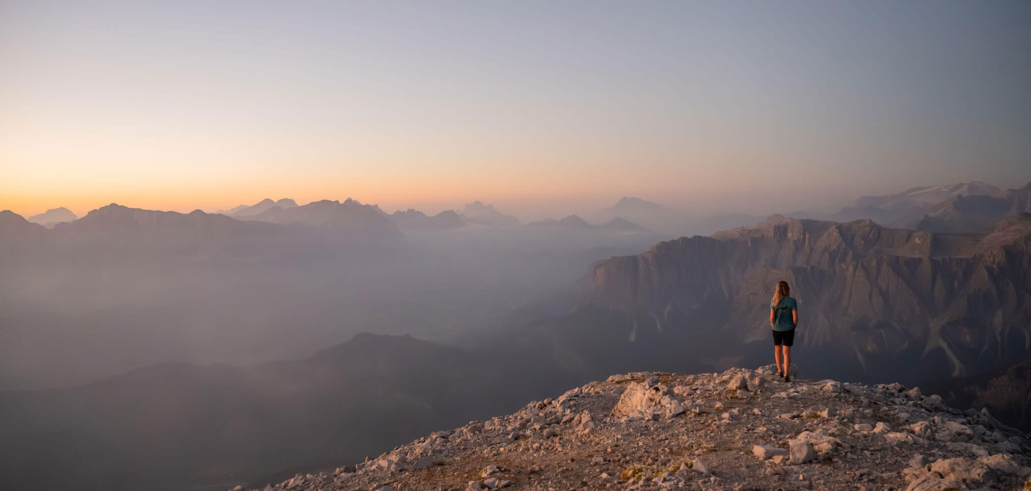 Eine Person steht bei Sonnenaufgang auf einem felsigen Berg und blickt auf nebelverhangene Gipfel und eine weite, dunstige Landschaft. - Hotel Oberwiesen