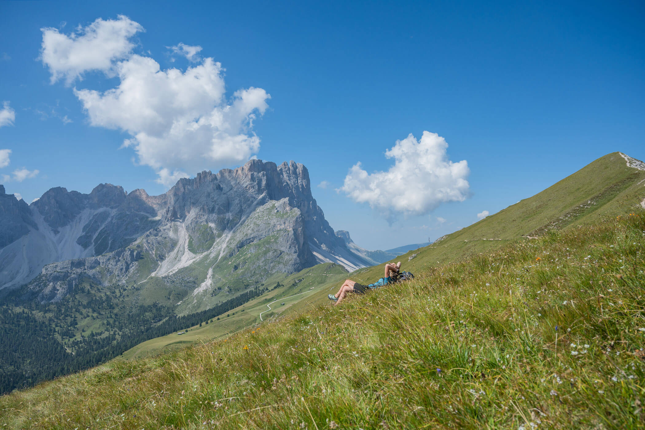 Eine Person liegt auf einem grasbewachsenen Hang, im Hintergrund sind Berggipfel und Wolken vor blauem Himmel zu sehen. - Hotel Oberwiesen