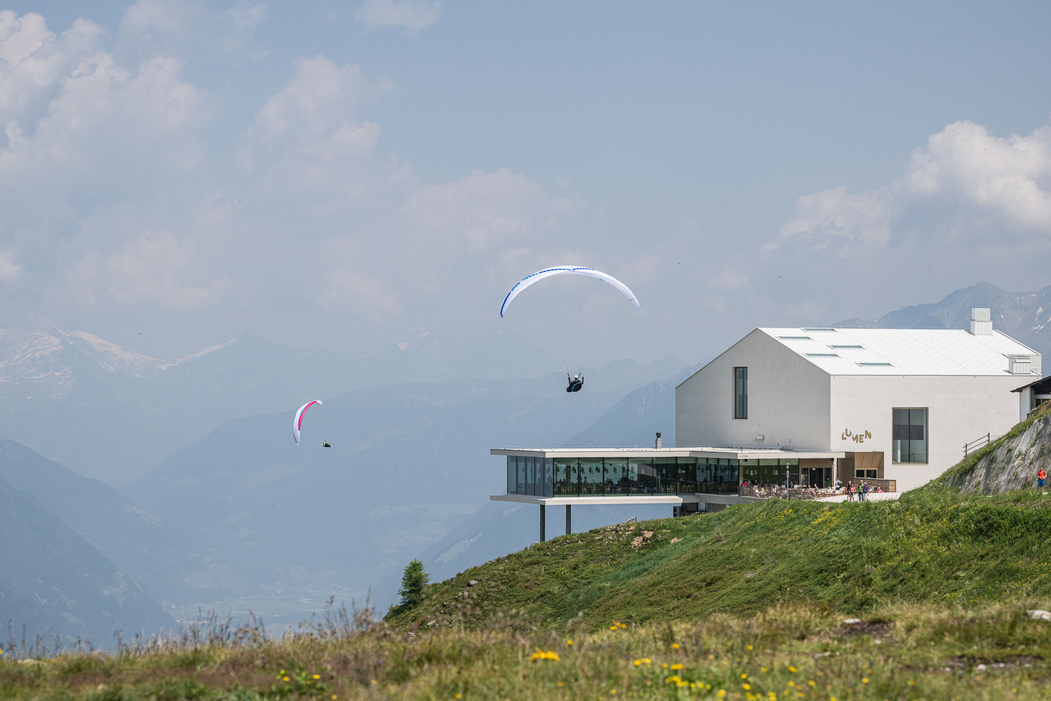 Ein modernes Gebäude am Berghang, über dem Gleitschirmflieger am Himmel über einem grünen Hang schweben. - Hotel Oberwiesen
