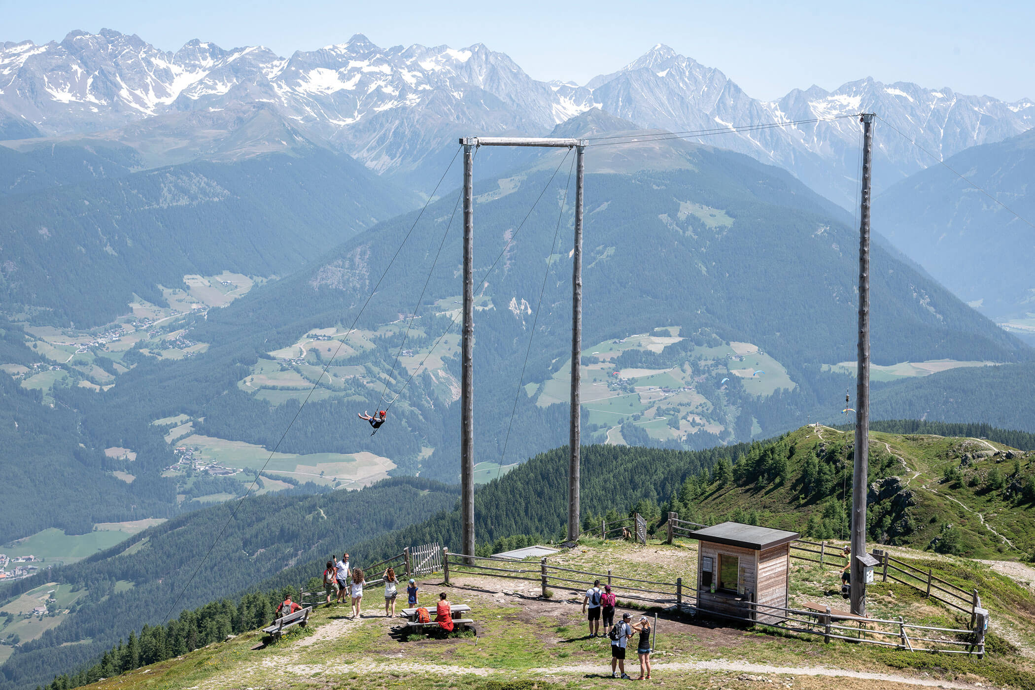 Eine Person schwingt sich hoch über einer Berglandschaft, während Menschen zuschauen und im Hintergrund schneebedeckte Gipfel zu sehen sind. - Hotel Oberwiesen