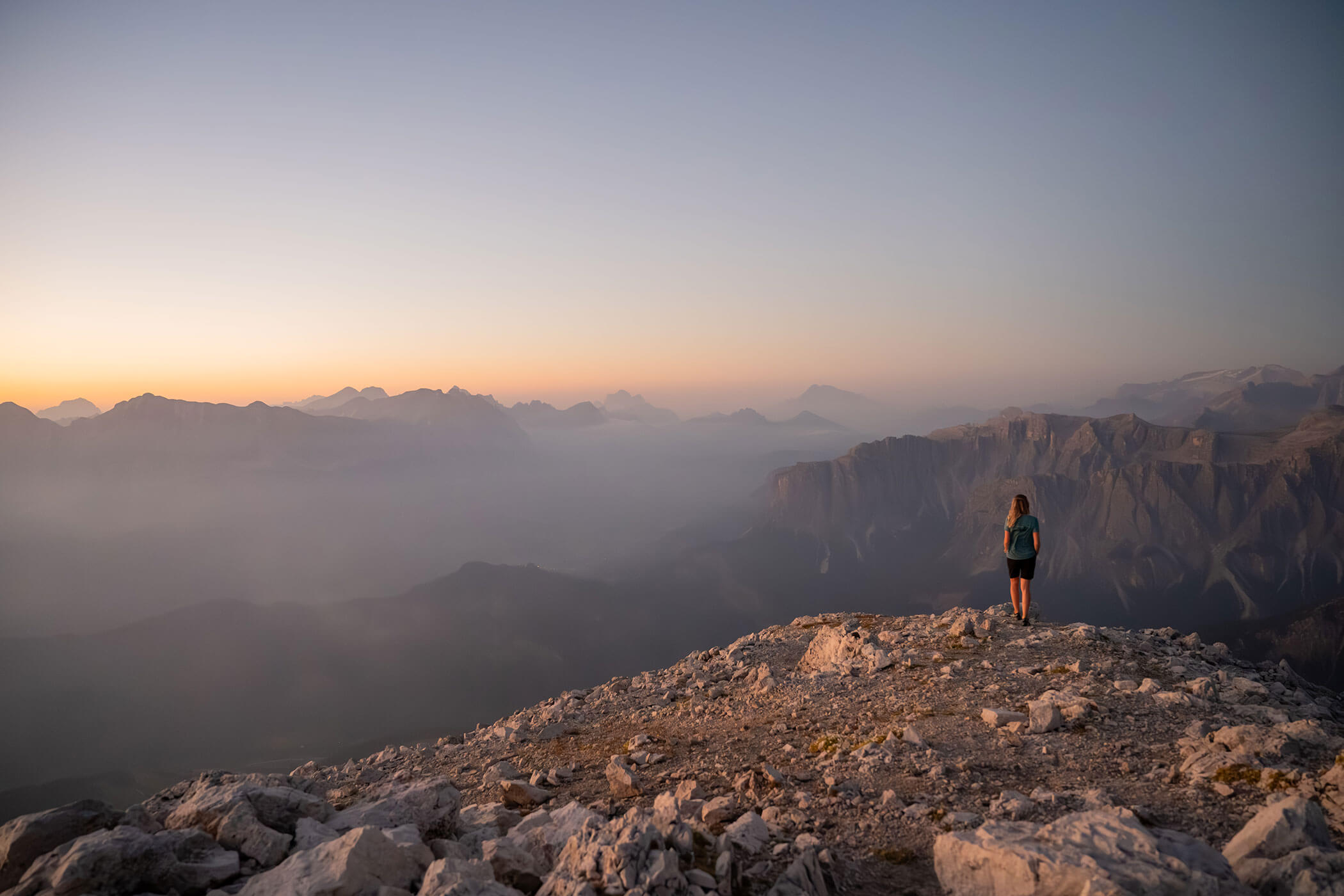 Eine Person steht bei Sonnenaufgang auf einem felsigen Berggipfel und blickt auf nebelverhangene Berge und einen dunstigen Himmel. - Hotel Oberwiesen