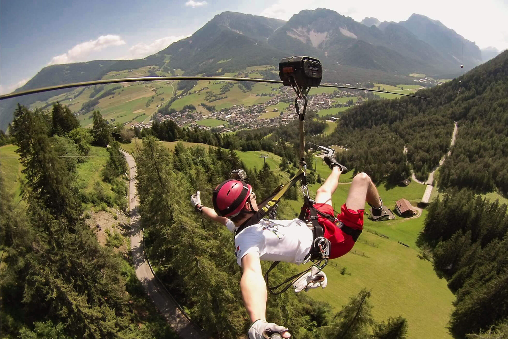 Eine Person, die auf einer Seilrutsche über ein üppig grünes Tal mit Bergen und Bäumen unter einem strahlend blauen Himmel gleitet. - Hotel Oberwiesen