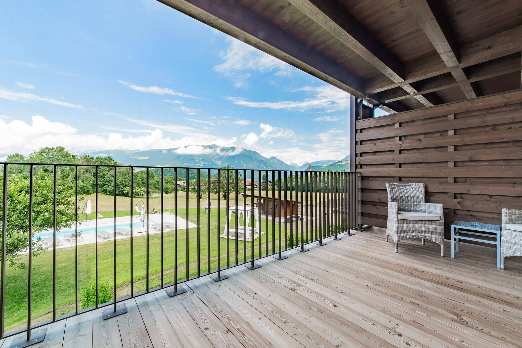 Ampio balcone in legno con sedie, vista sulle montagne e una piscina in un giardino con prato sotto un cielo azzurro. - Hotel Oberwiesen