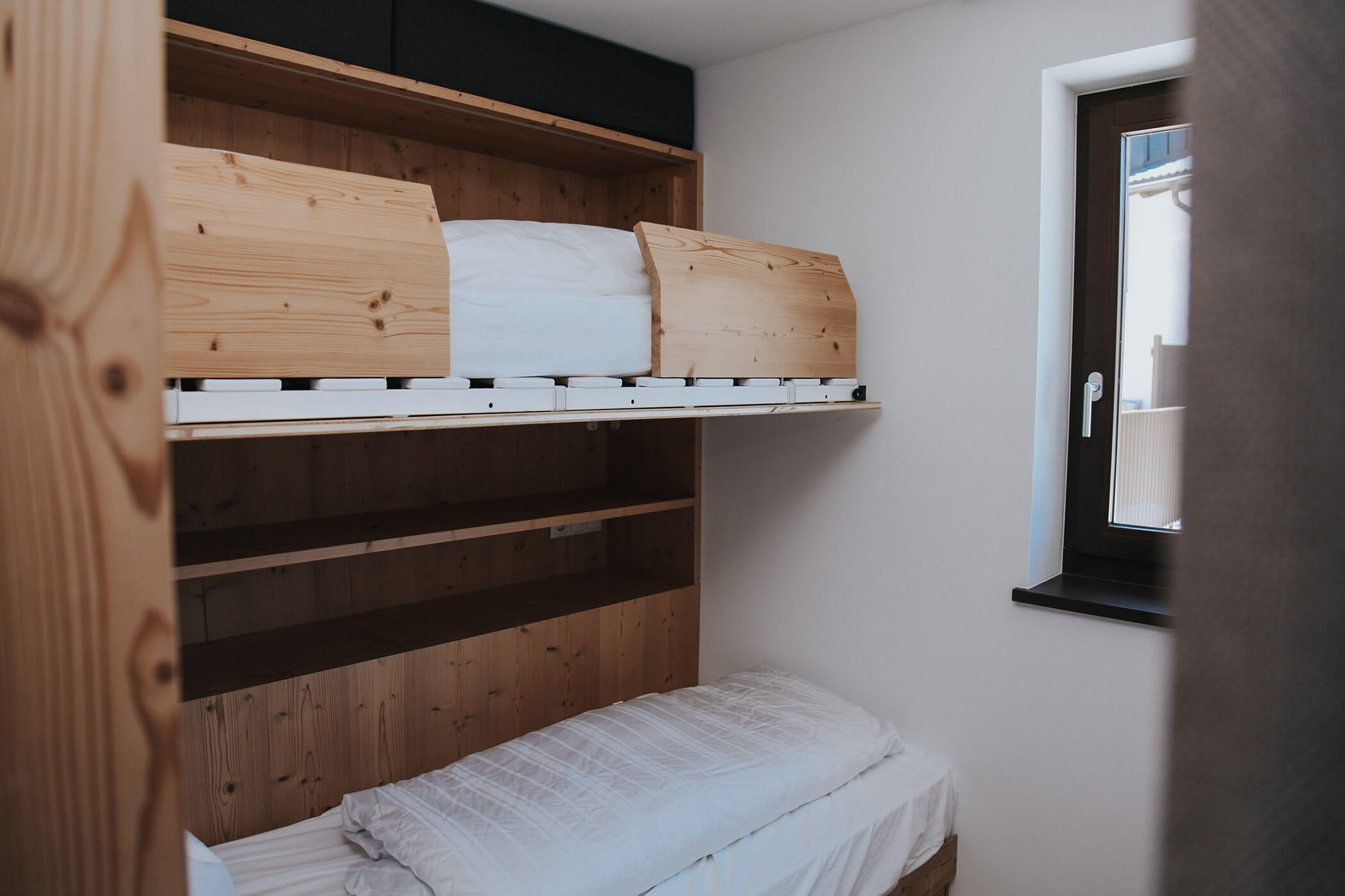 Piccola stanza con letti a castello in legno, biancheria da letto bianca, mensole e una finestra con infisso nero. - Hotel Oberwiesen