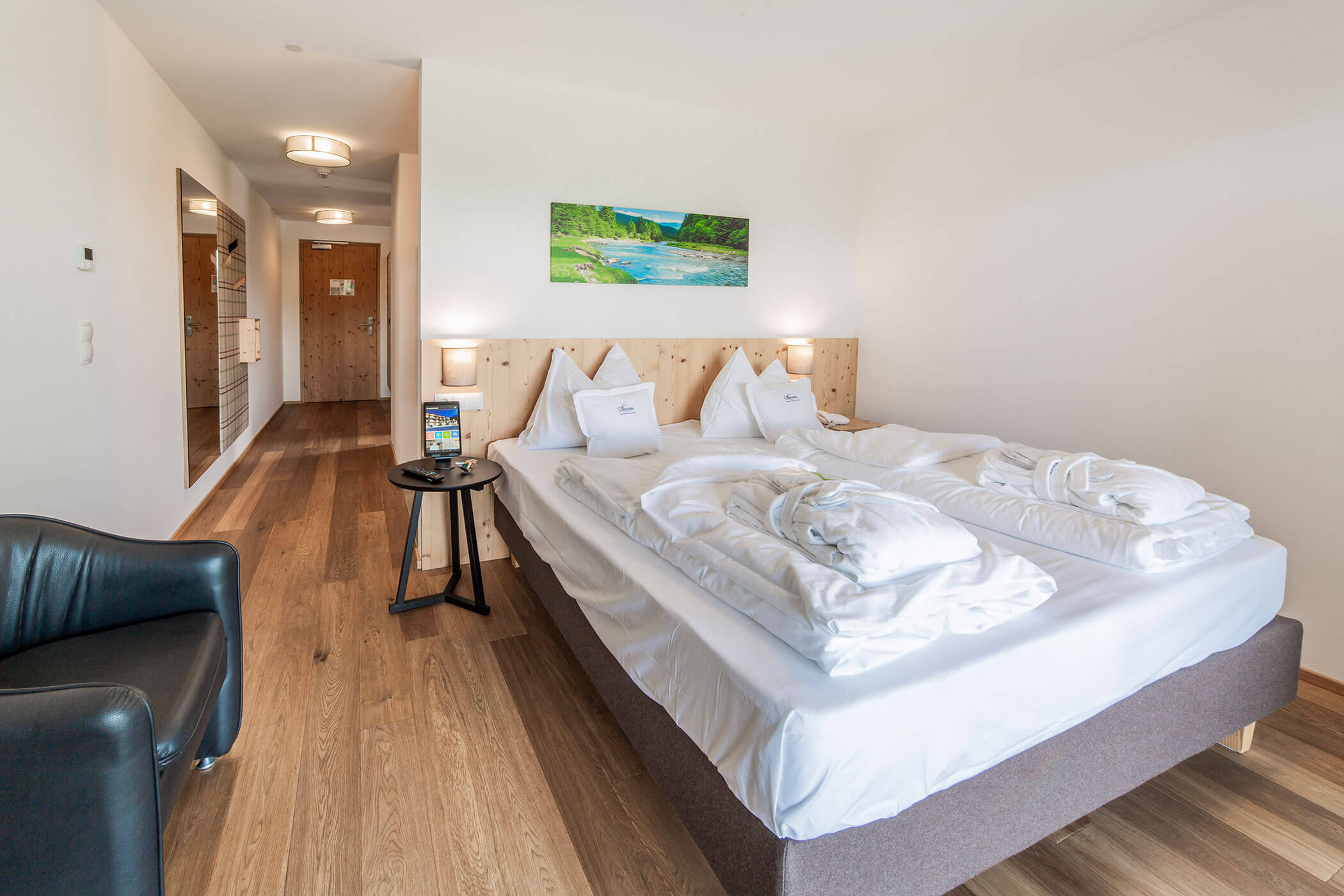 Camera d'albergo luminosa con letto matrimoniale, accappatoi, lampade, una sedia nera e un quadro appeso sopra la testiera del letto. - Hotel Oberwiesen