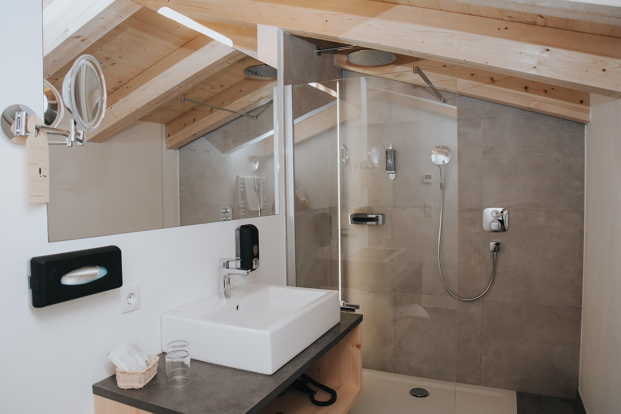 Bagno moderno con lavabo, specchio, box doccia in vetro e soffitto inclinato in legno. - Hotel Oberwiesen