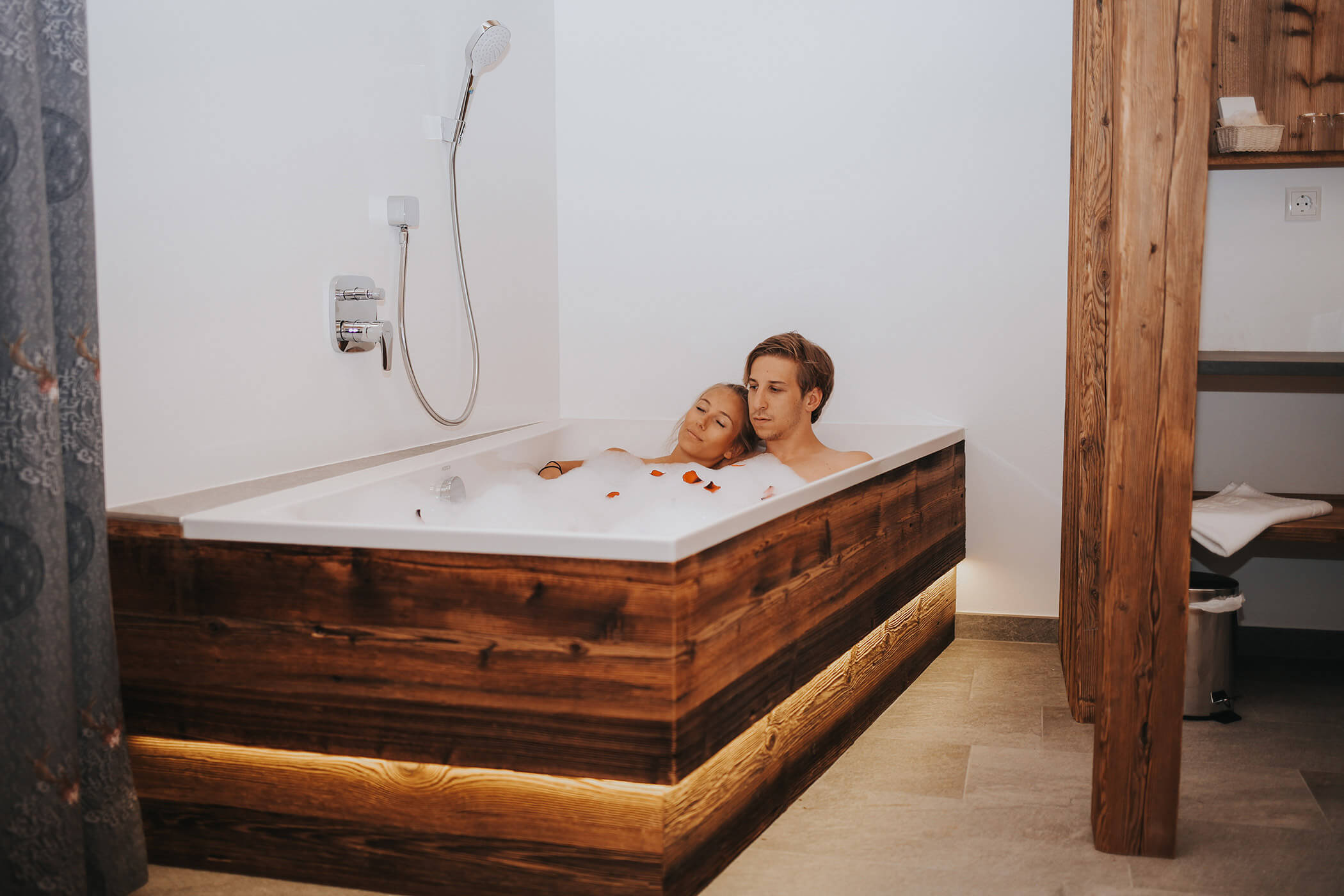 Coppia che si rilassa insieme in una vasca da bagno in legno con bolle e petali di rosa in un bagno accogliente. - Hotel Oberwiesen