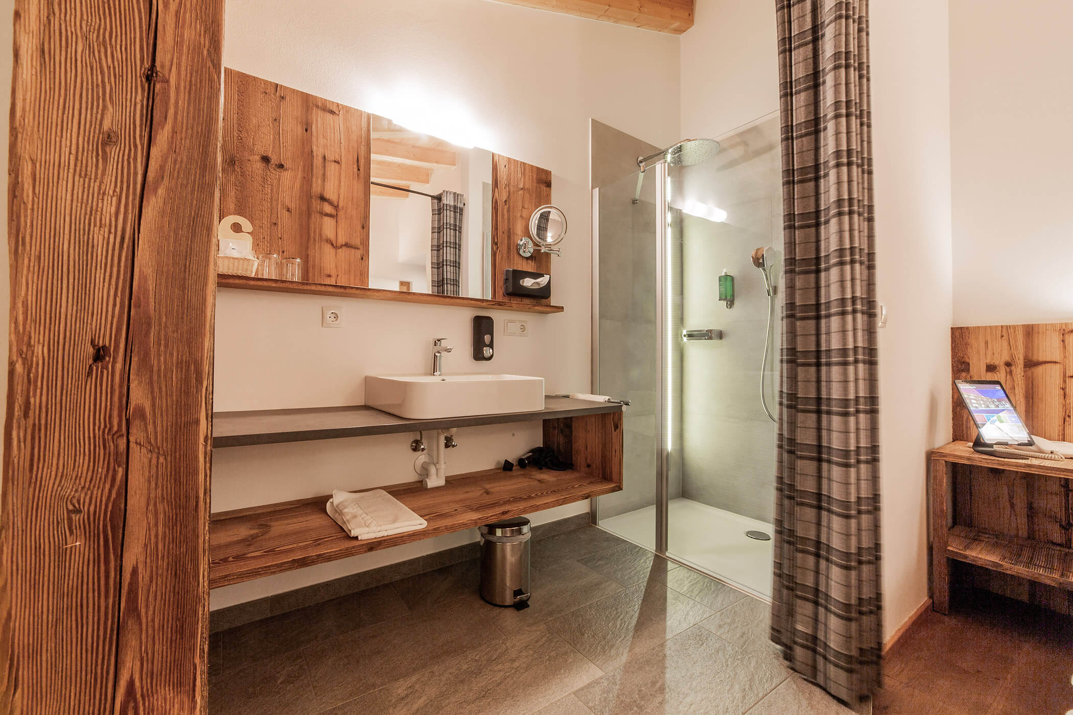 Bagno moderno con dettagli in legno, lavandino, specchio, doccia e un asciugamano su una mensola sotto il lavandino. - Hotel Oberwiesen