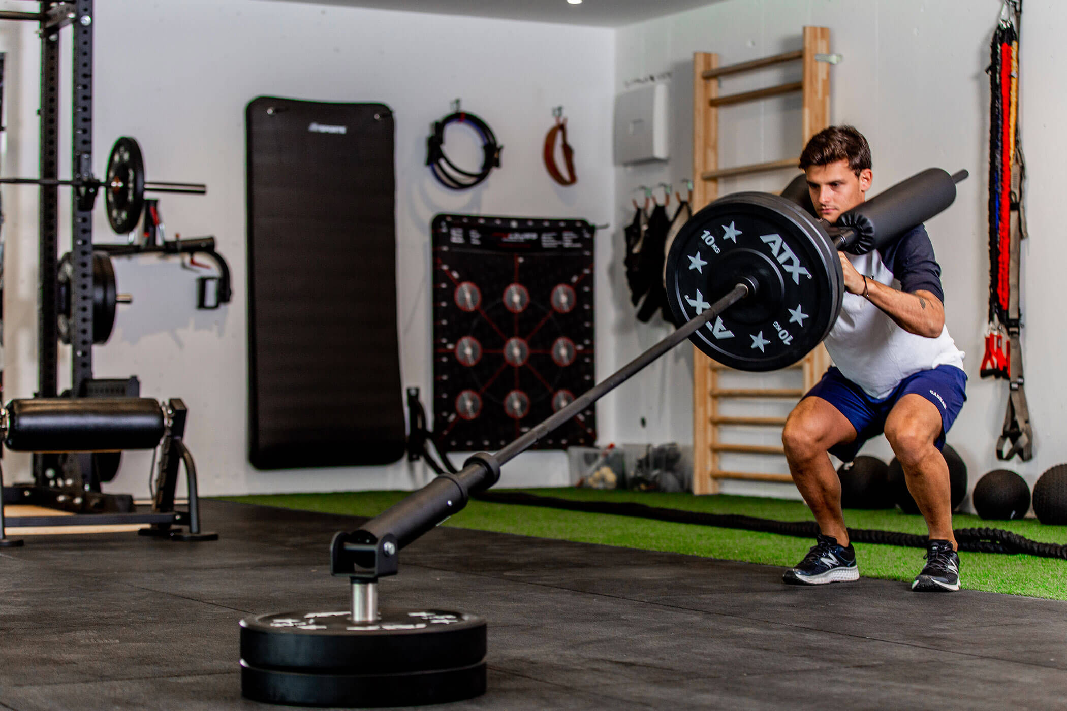 Ein Mann in Turnhose und T-Shirt führt in einem Fitnessstudio eine Landmine-Kniebeuge mit einer Langhantel aus. - Hotel Oberwiesen