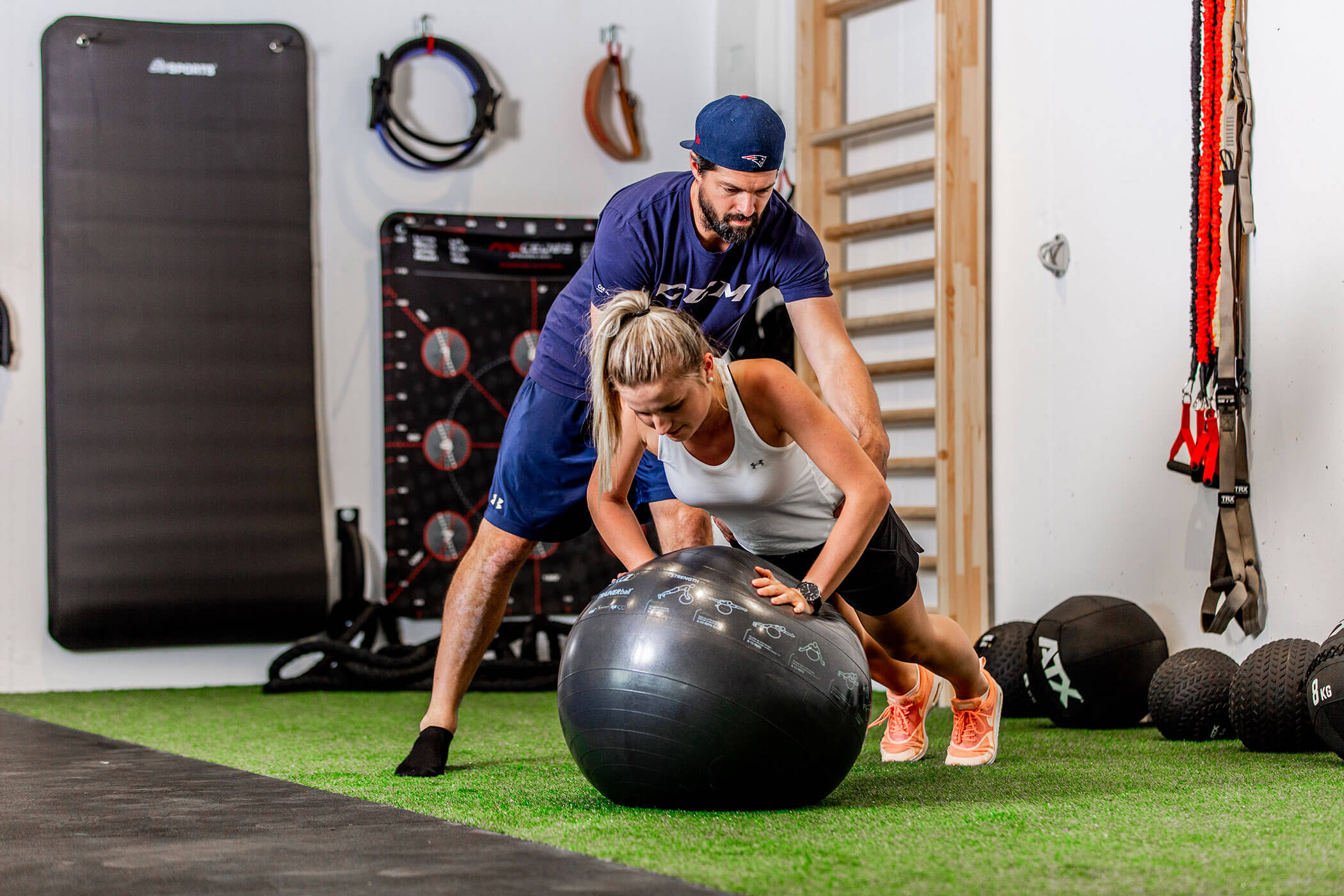 Ein Mann hilft einer Frau, die auf einem Gymnastikball in einem Fitnessstudio mit Trainingsgeräten Liegestütze macht. - Hotel Oberwiesen