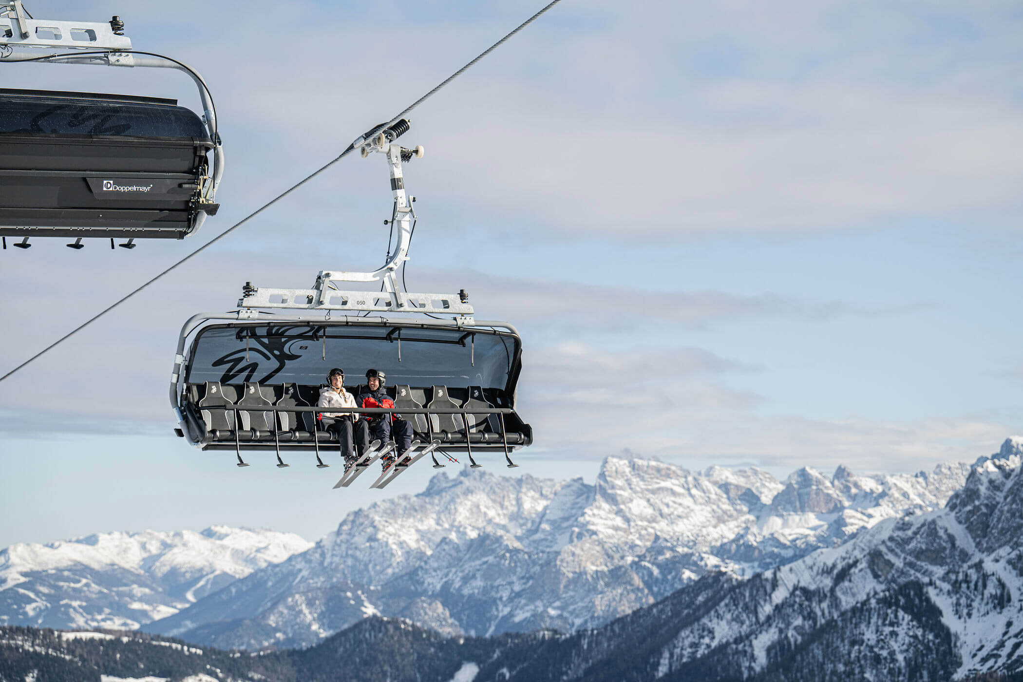 Drei Menschen fahren mit einem Skilift über verschneite Berge unter einem bewölkten Himmel. - Hotel Oberwiesen